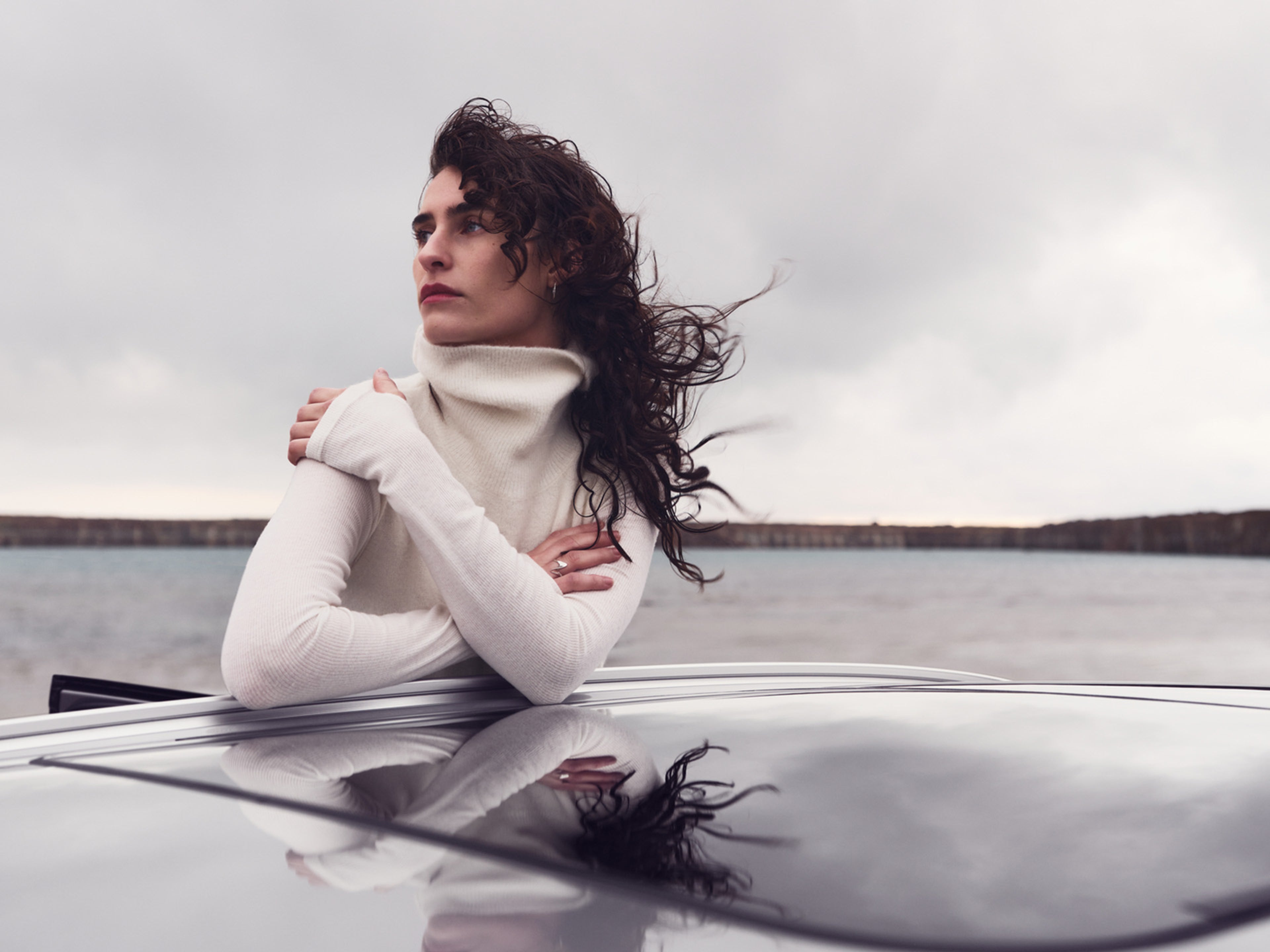 Mulher morena vestida de branco inclinada no teto panoramico de um volvo, com o mar nas costas