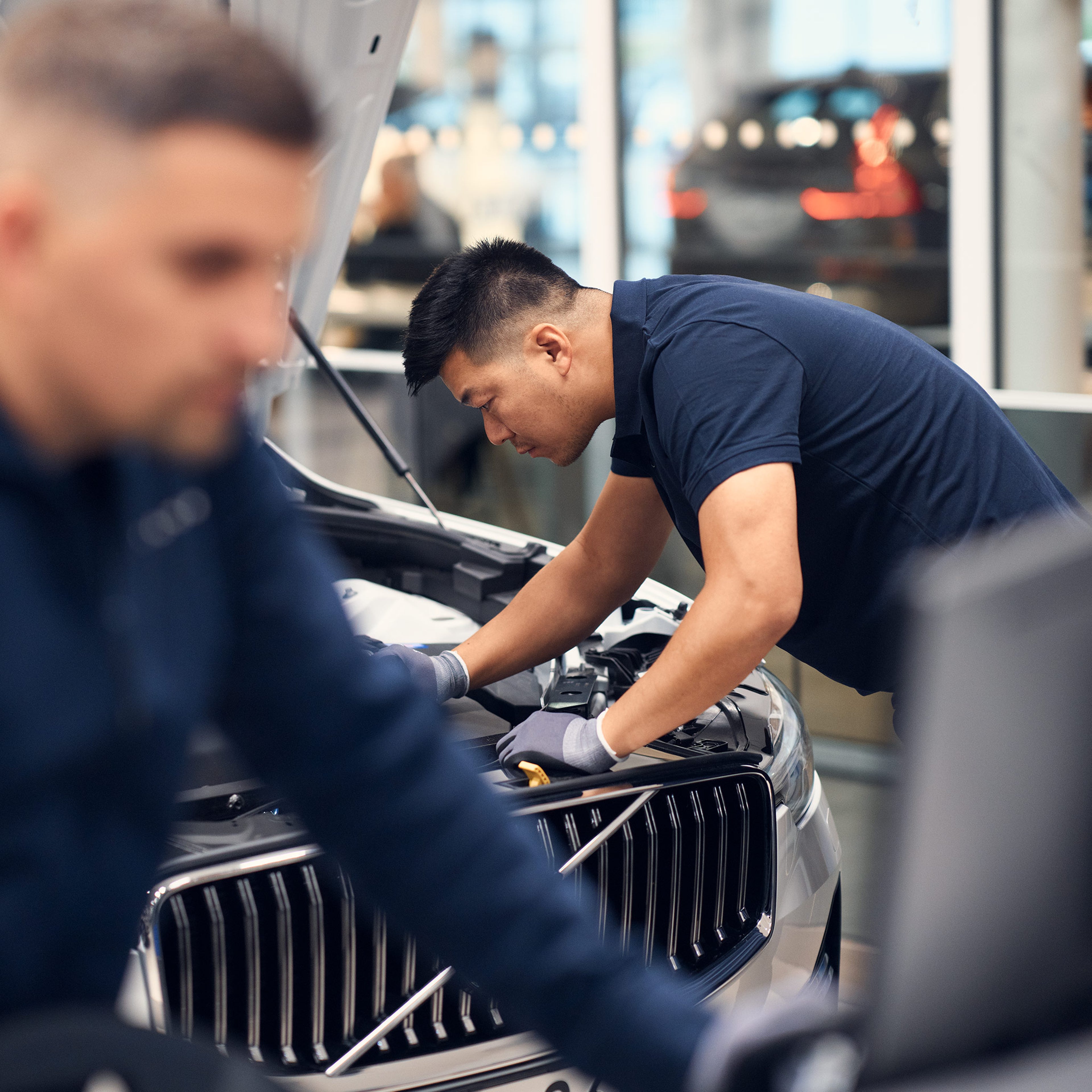 Volvo Omamekaanikko tutkii Volvo auton moottoritilaa. Etualalla toinen Volvo Omamekaanikko katsoo tietokoneen näyttöä työasemalla.