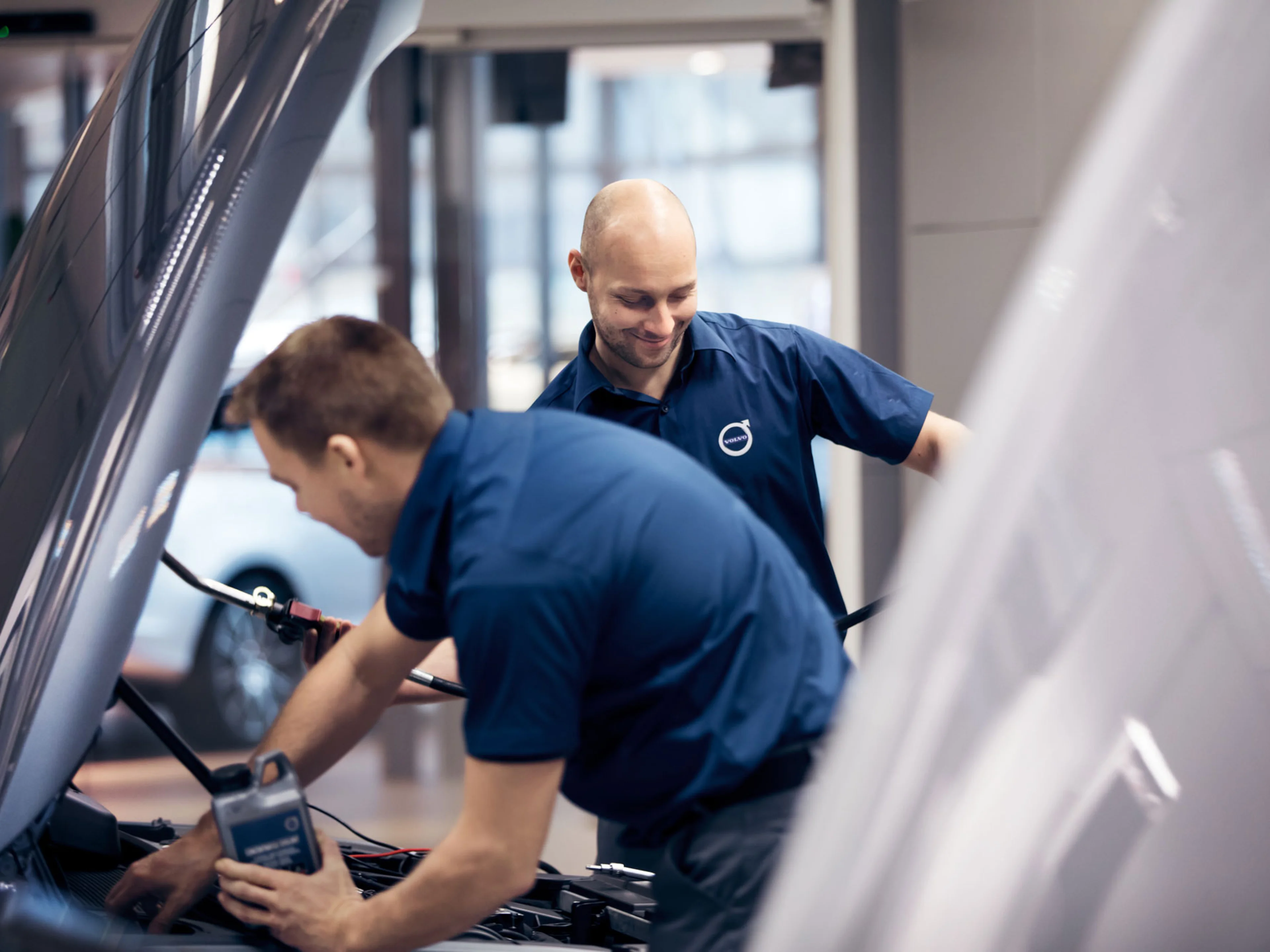 To Volvo mekanikere i blå uniformer arbejder på en bilmotor med åben motorhjelm indendørs. De er engagerede og fokuserede, hvilket giver en professionel tone.