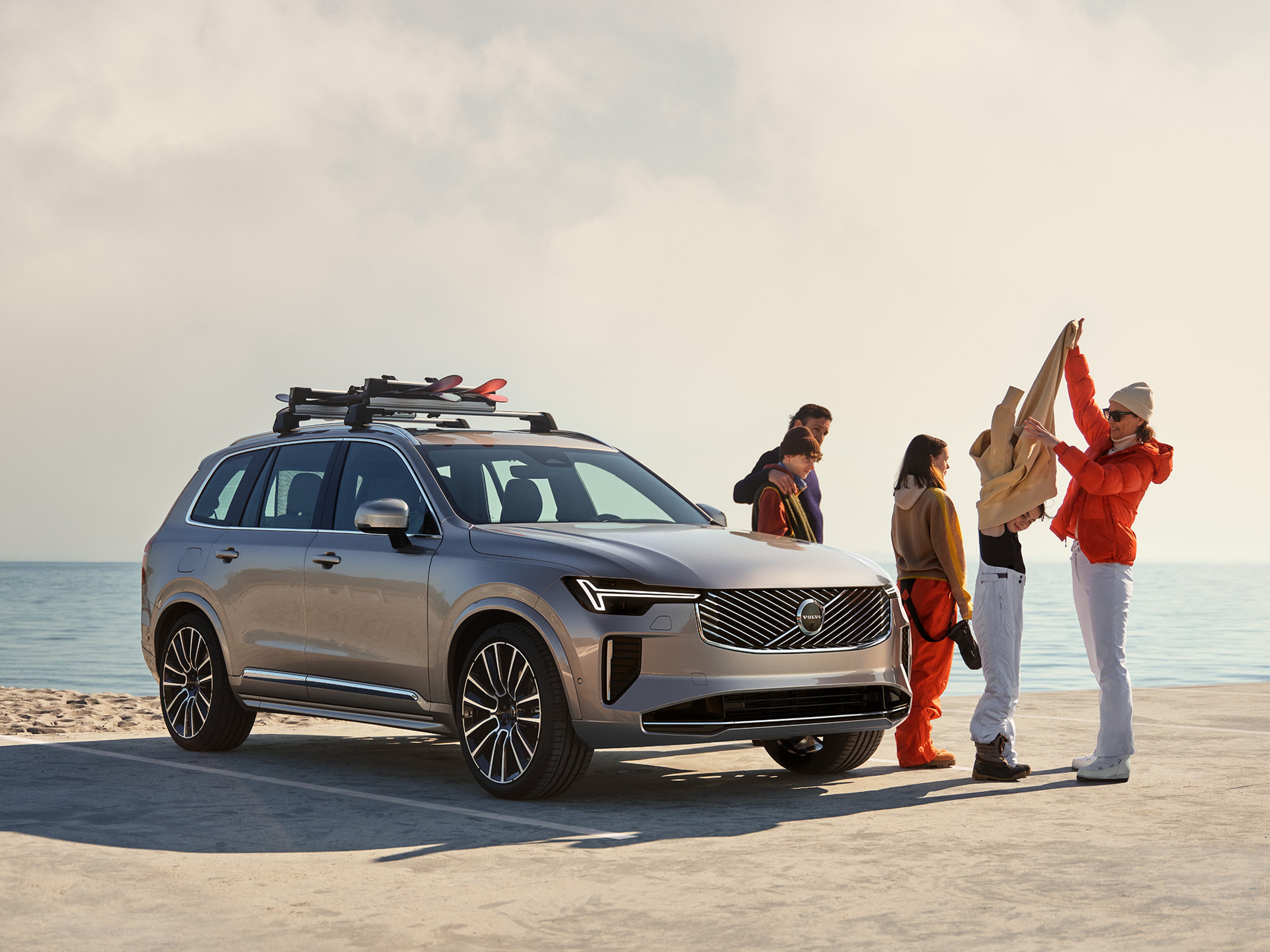 Viiden hengen perhe riisuu talvivaatteita hiekkarannan viereen parkkeeratun Bright Dusk -värisen Volvo XC90 lataushybridiauton vieressä.
