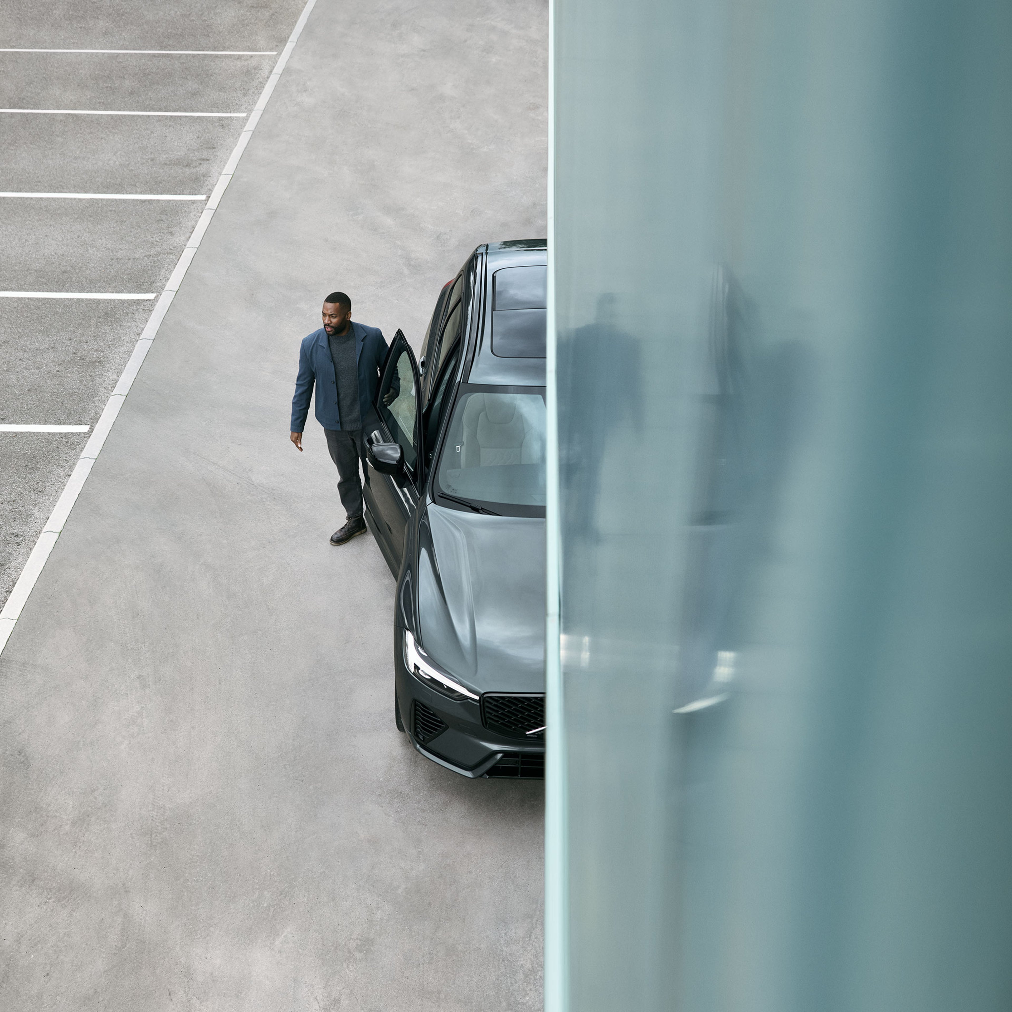 Mies seisoo parkkipaikalla Forest lake -värisen Volvo XC60 lataushybridikatumaasturin vieressä. Kuvaa halkoo turkoosi seinä, jonka pinnasta näkyy heijastuskuvana pysäköity auto ja mies.
