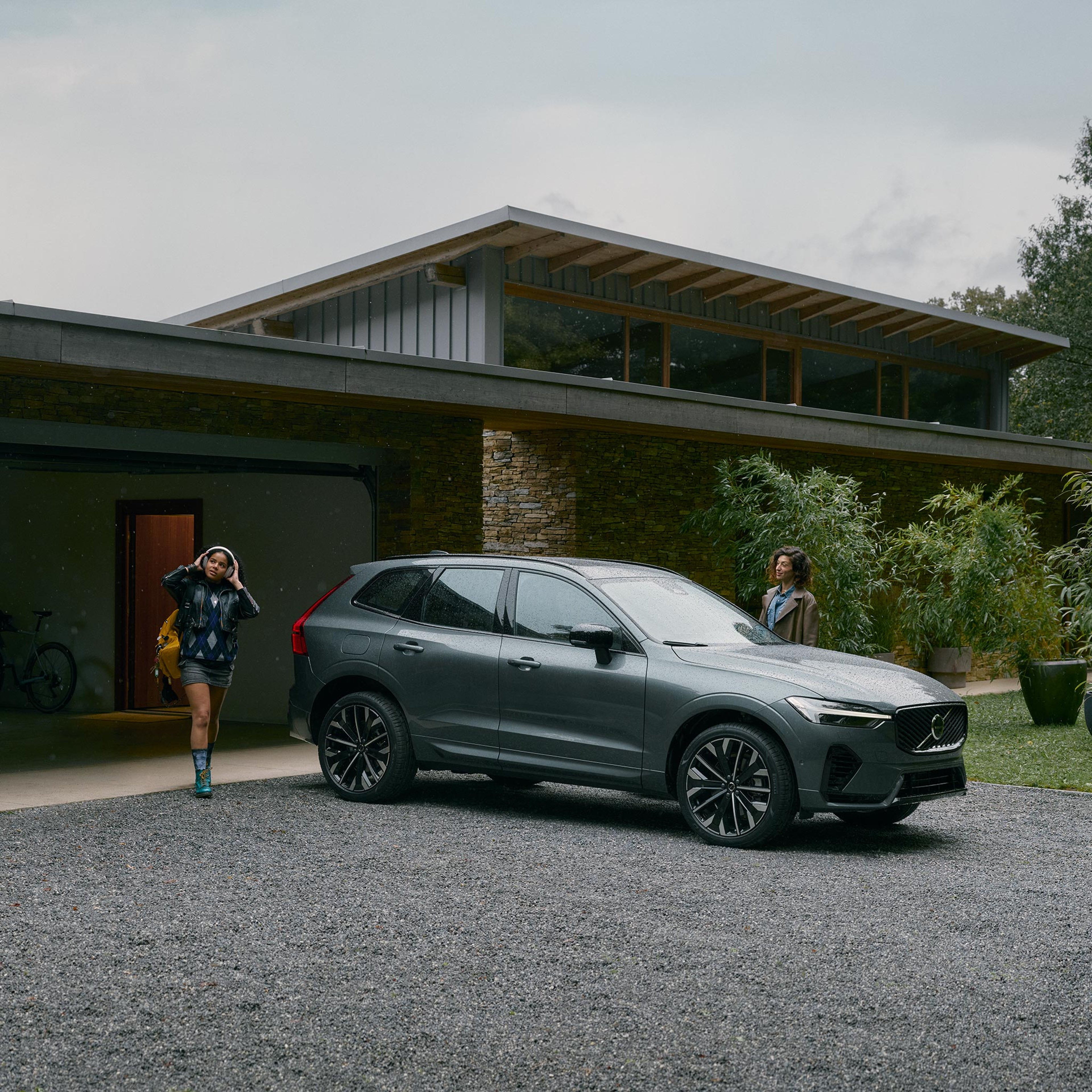 Tyttö kävelee kohti Forest lake -väristä Volvo XC60 lataushybridikatumaasturia modernin liuskekivipintaisen omakotitalon pihassa. Nainen seisoo kuljettajan puoleisen oven vieressä ja odottaa tyttöä.