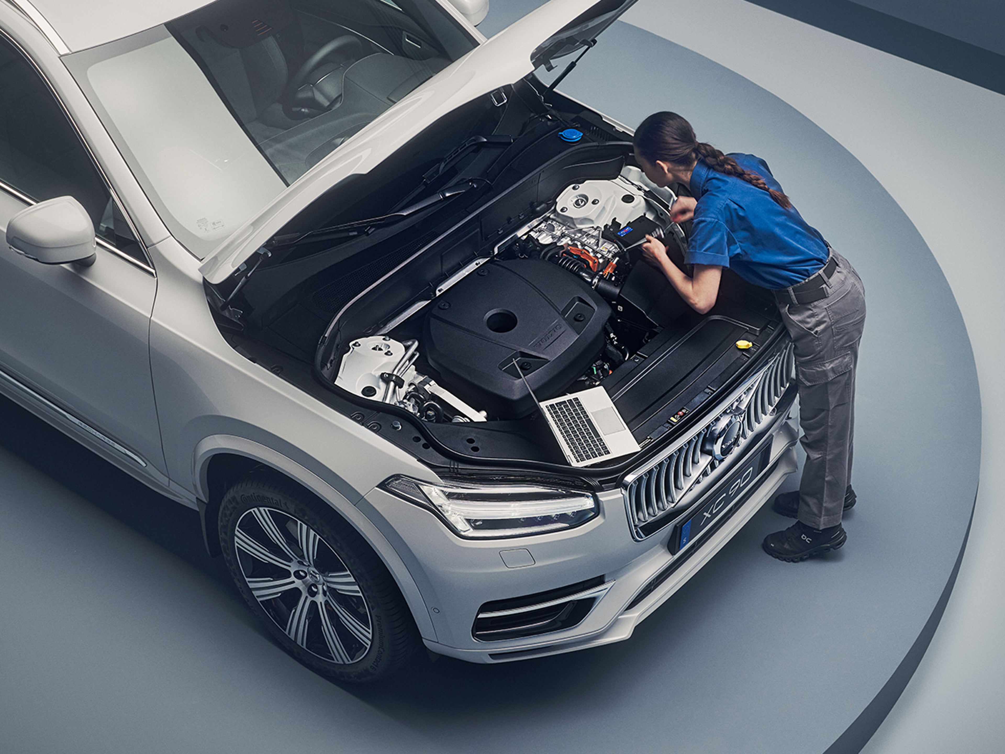 A Volvo mechanic inspects the engine of a silver Volvo SUV with a laptop beside her, showcasing automotive maintenance in a modern setting.