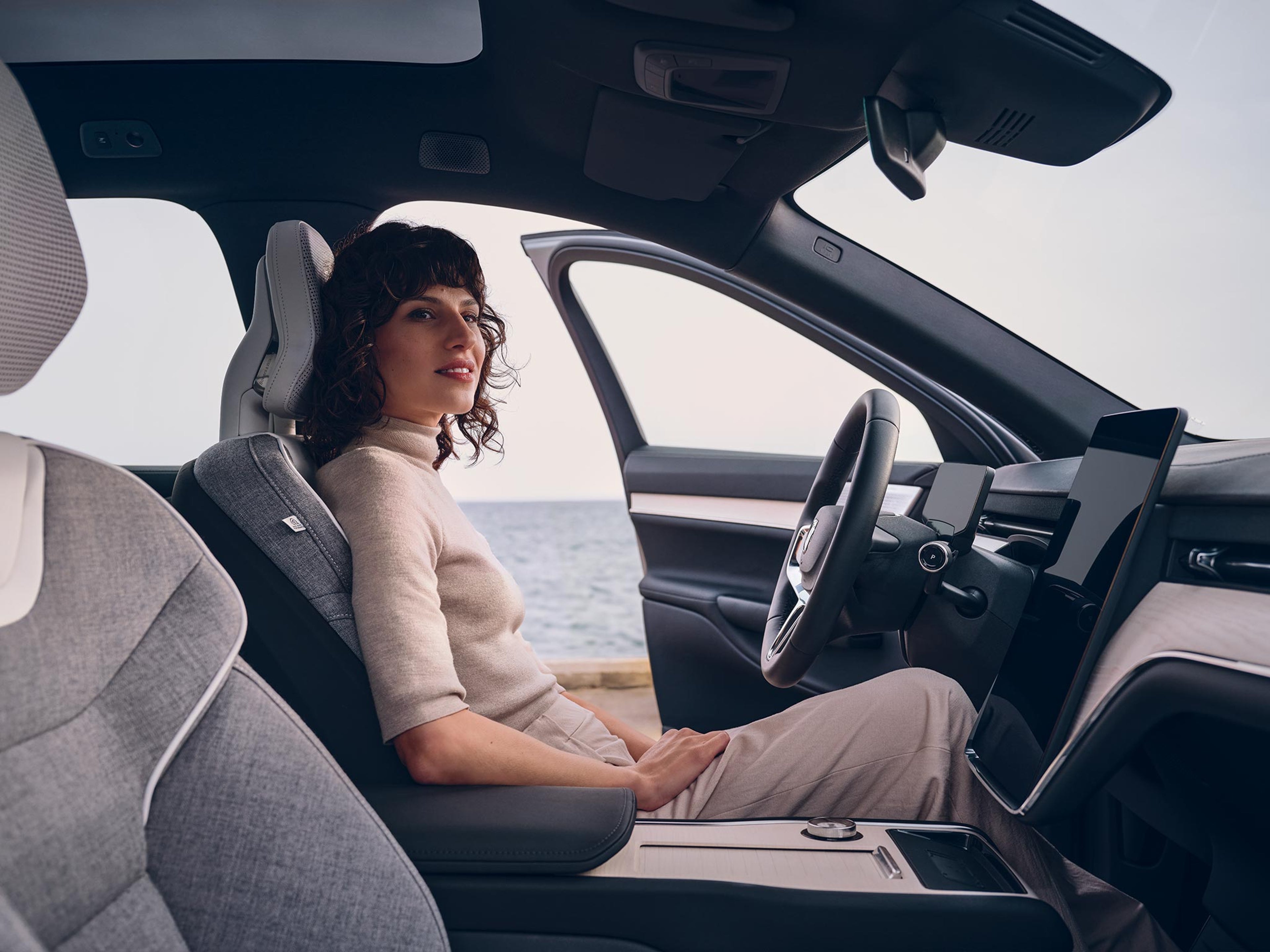 Nainen istuu Volvo EX90 täyssähköauton kuskin paikalla ja auton etuovi on auki, taustalla näkyy meri.