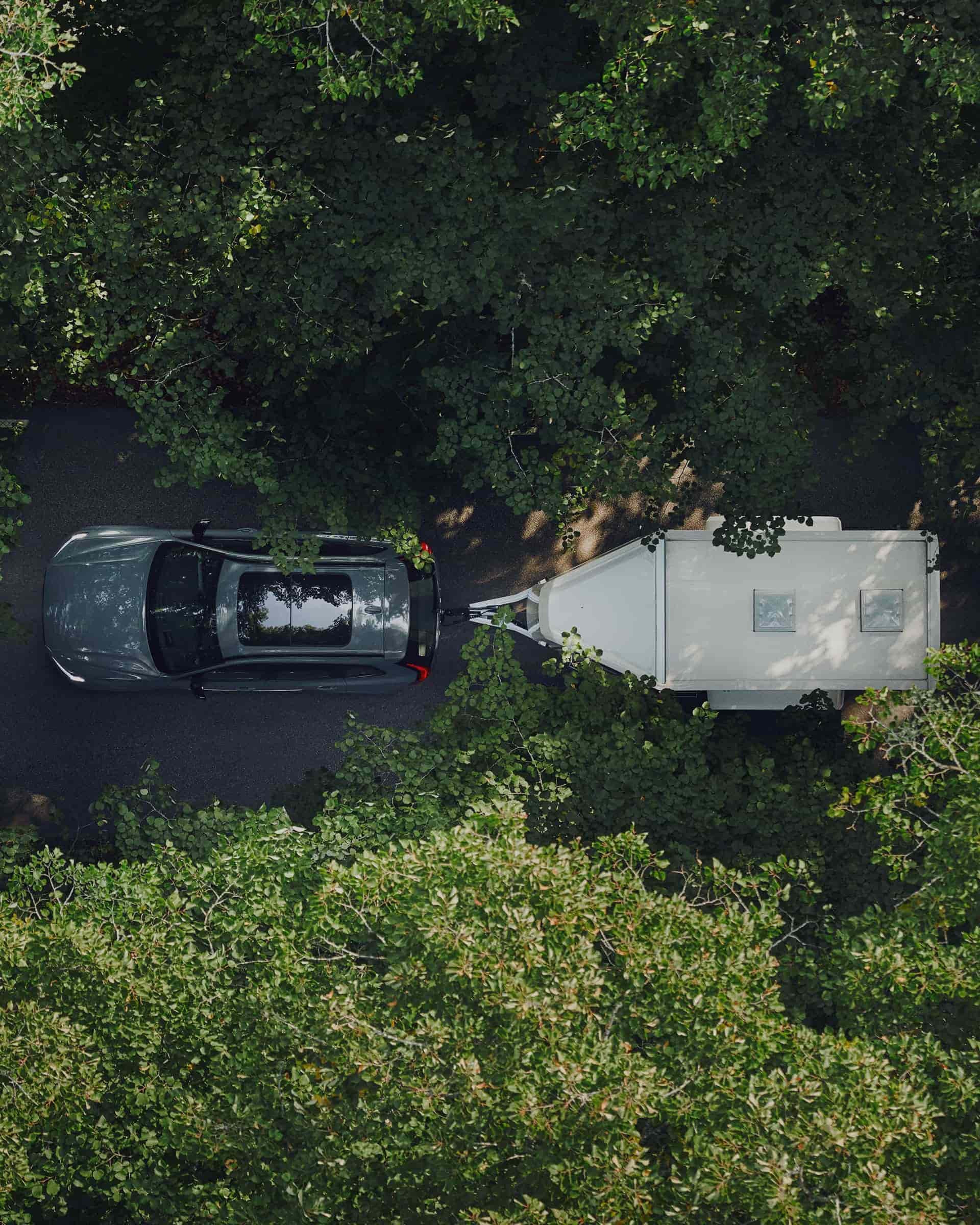 Bovenaanzicht van een grijze Volvo V90 met een aanhangwagen, rijdend op een weg door het bos."