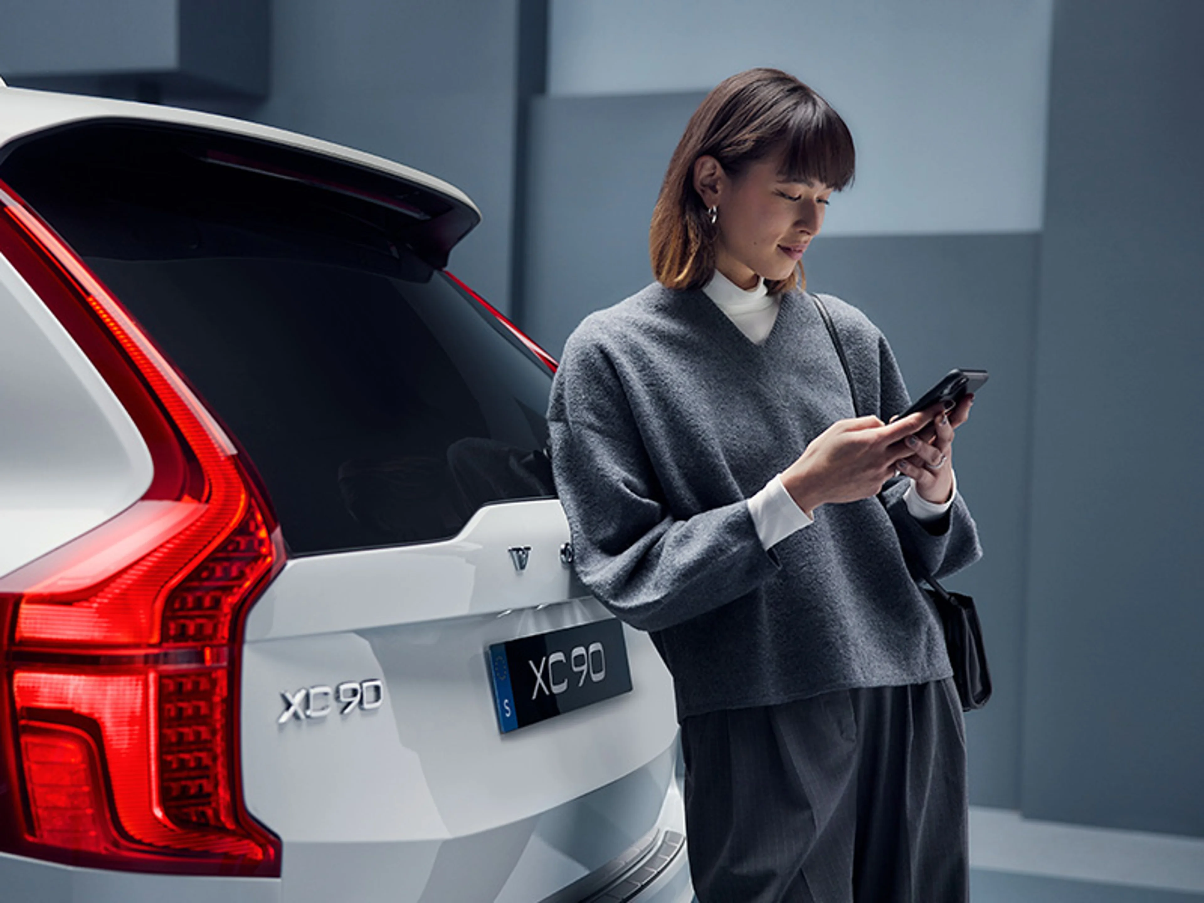 Une femme utilise son téléphone portable, appuyée contre l'arrière d'une Volvo XC90 blanche.