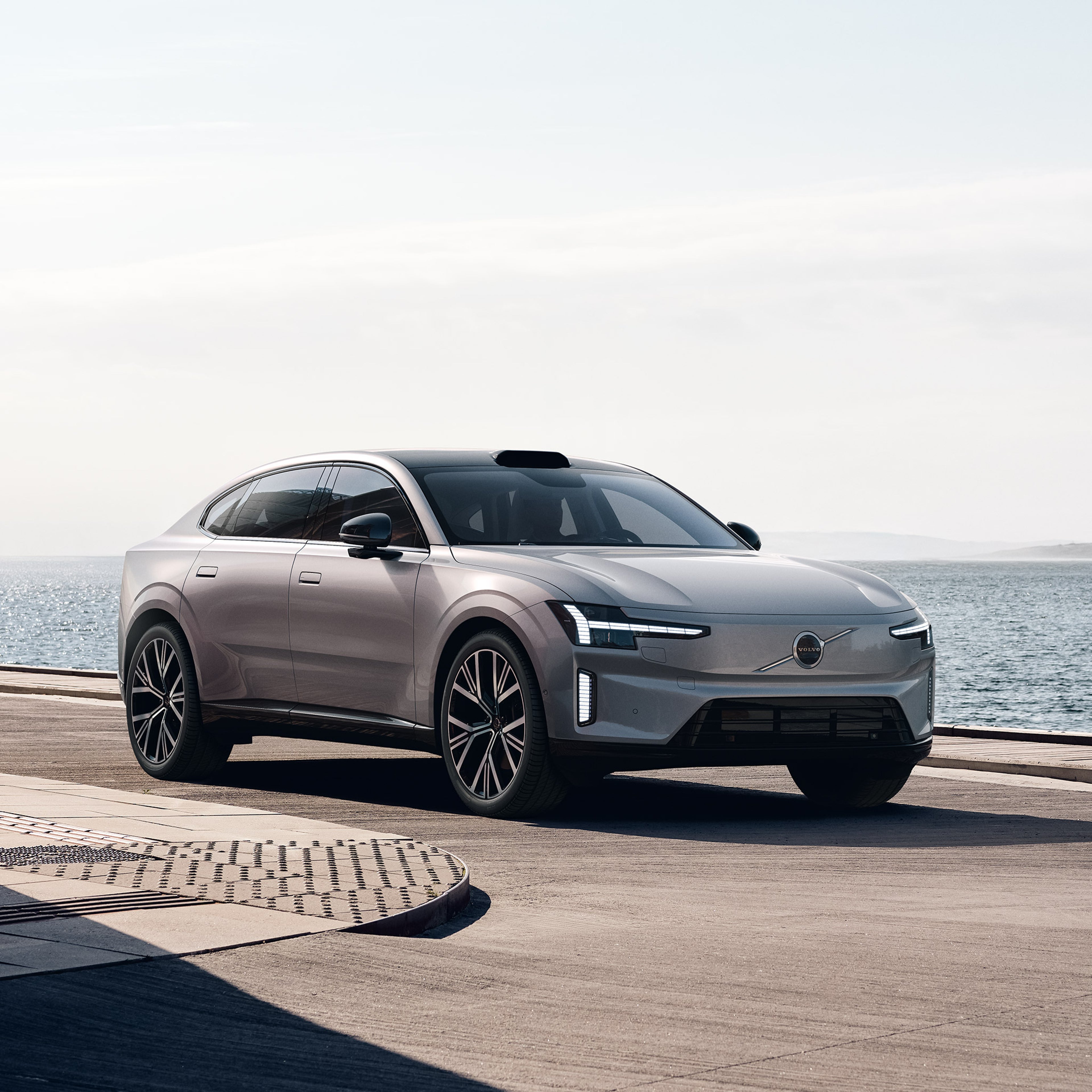 Aurora Silver -värinen Volvo ES90 täyssähköinen sedan pysäköitynä rantaan kivetykselle. Taustalla meri ja vaalea taivas.