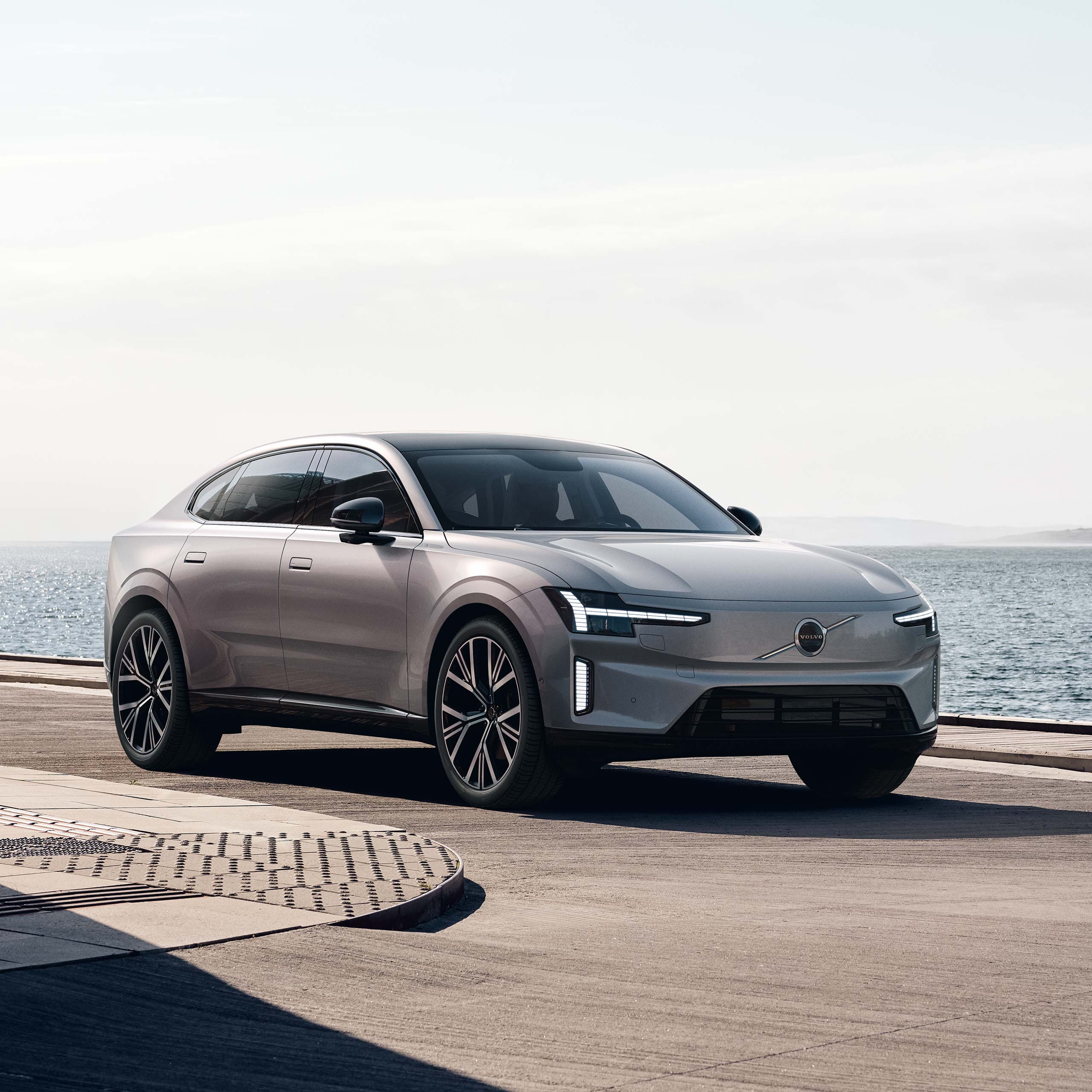 Aurora Silver -värinen Volvo ES90 täyssähköinen sedan pysäköitynä rantaan kivetykselle. Taustalla meri ja vaalea taivas.