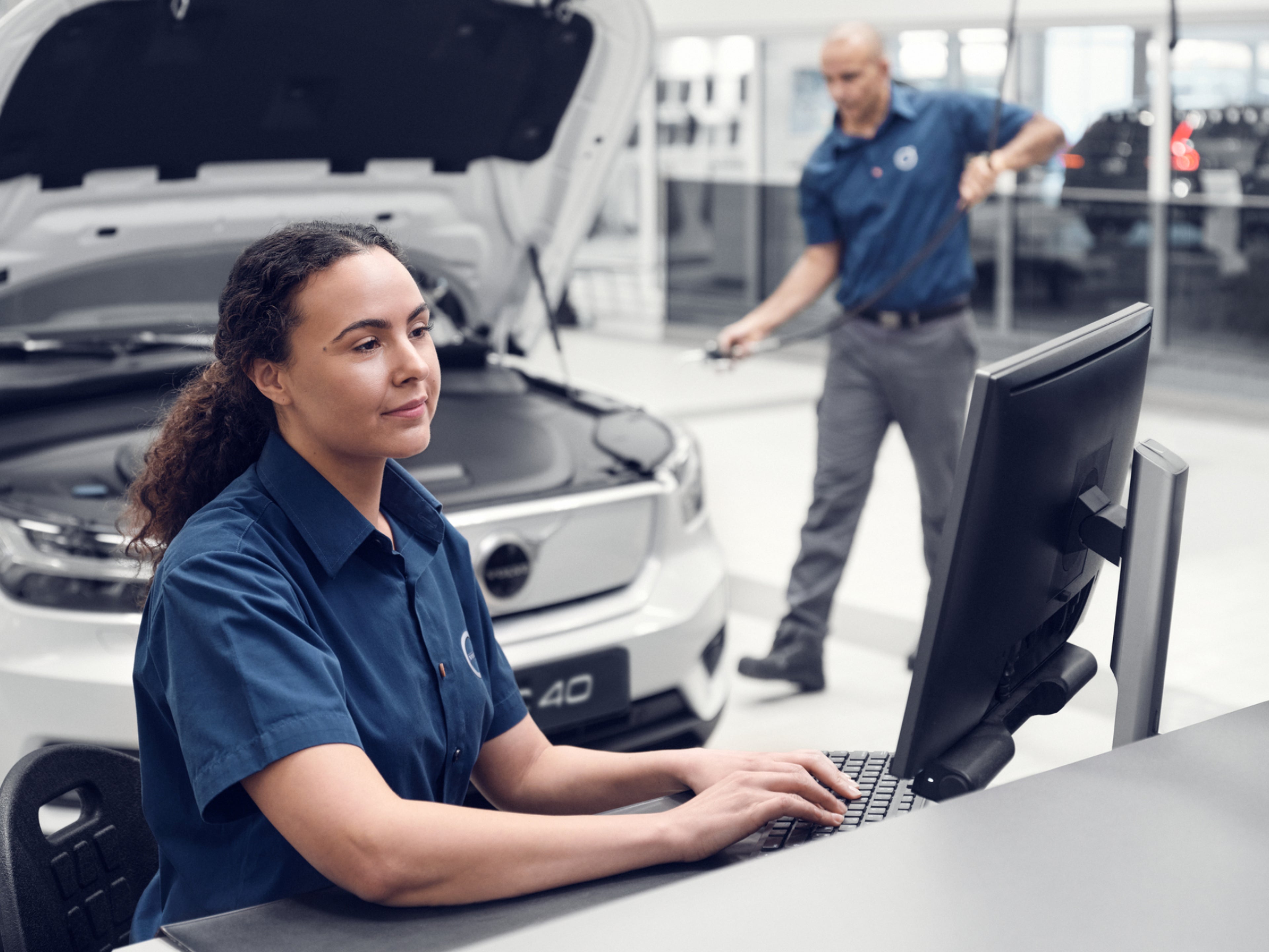 Eine weibliche Volvo Service Technikerin sitzt vor einem Analyse-Screen in einer Werkstatt, während ein männlicher Techniker einen Volvo EX40 inspiziert.