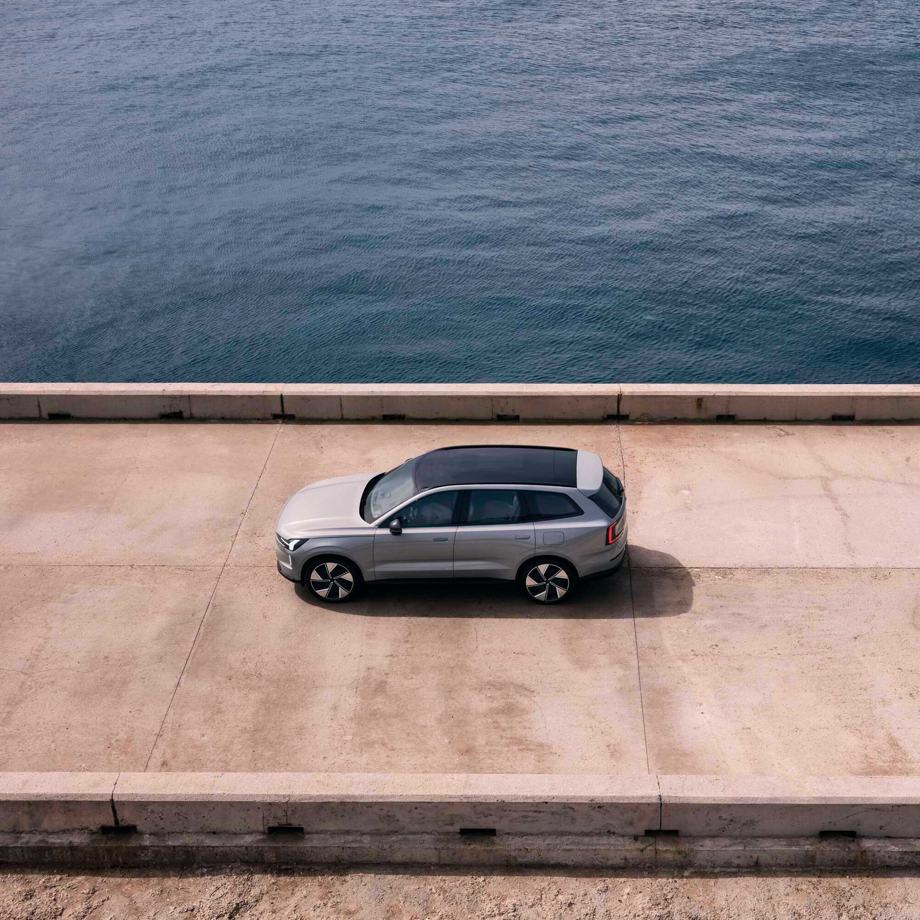 Sølvfarvet SUV parkeret på en betonmole ved et roligt, blåt hav – set oppefra. Scenen udstråler ro og frihed ved vandet.