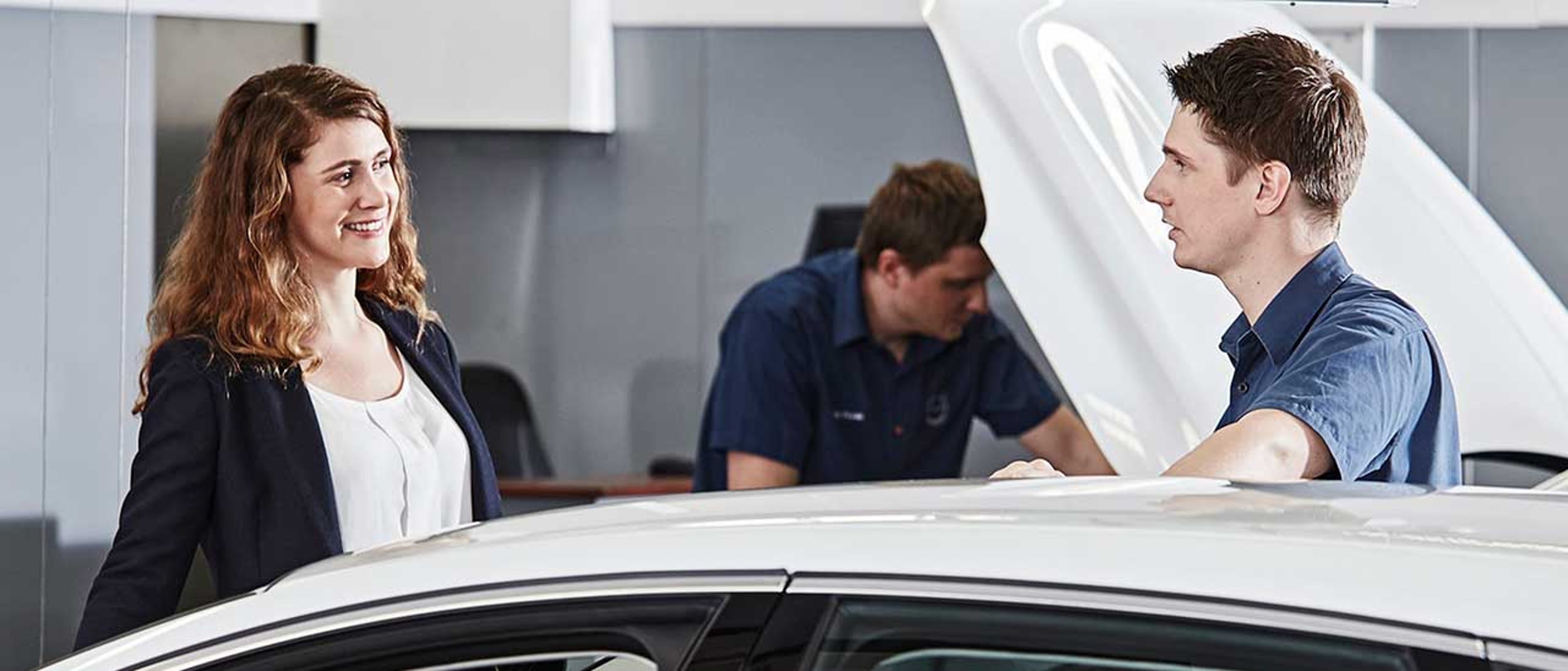 En kvinde smiler til en mand, der læner sig op ad en bil med åben motorhjelm af en Volvo bil på et Volvo servicecenter. En anden mekaniker arbejder i baggrunden. Atmosfæren er venlig.