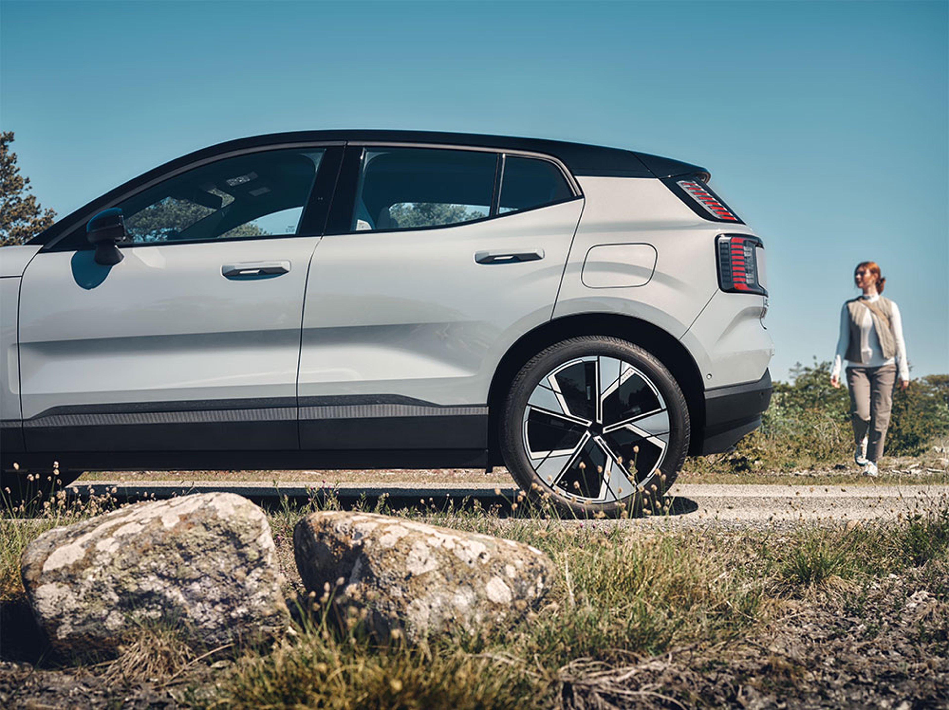 SUV EX30 Bleu Nuage garé en bordure d'un chemin de gravier dans un paysage ouvert sous un ciel bleu, une femme approche par le côté arrière