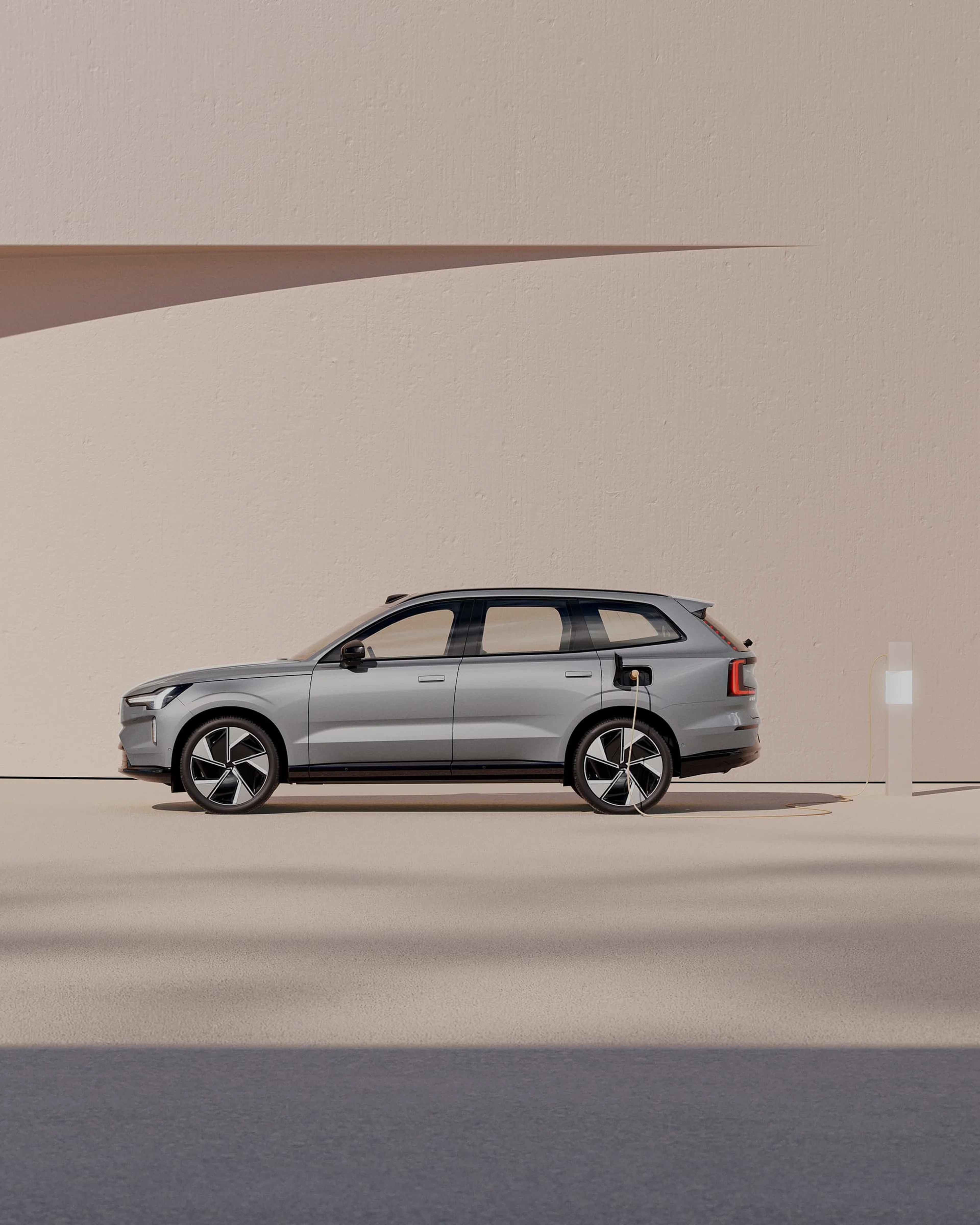 Vue latérale d'un Volvo EX90 gris à une station de charge sur fond orange pastel.