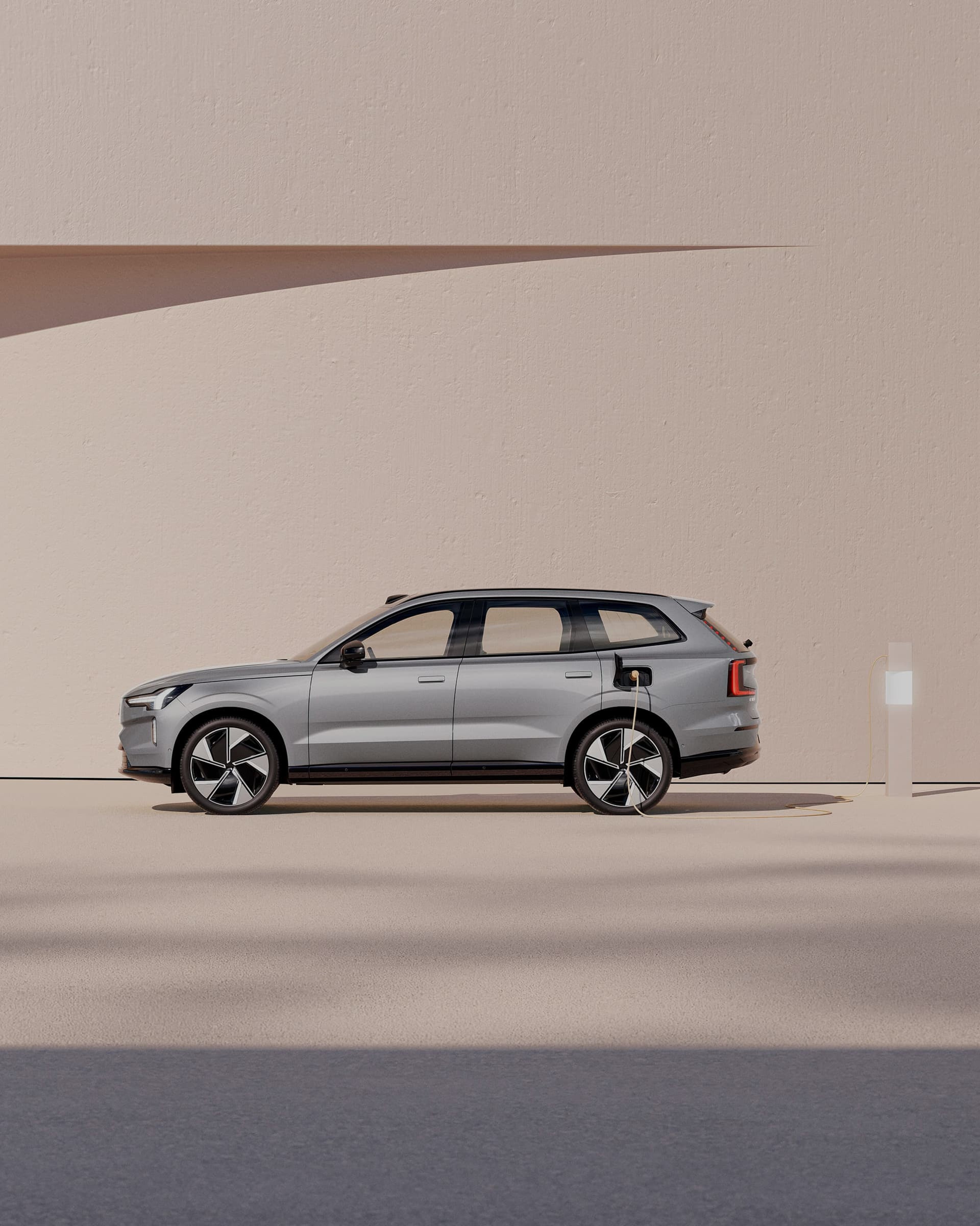 Vue latérale d'un Volvo EX90 gris à une station de charge sur fond orange pastel.