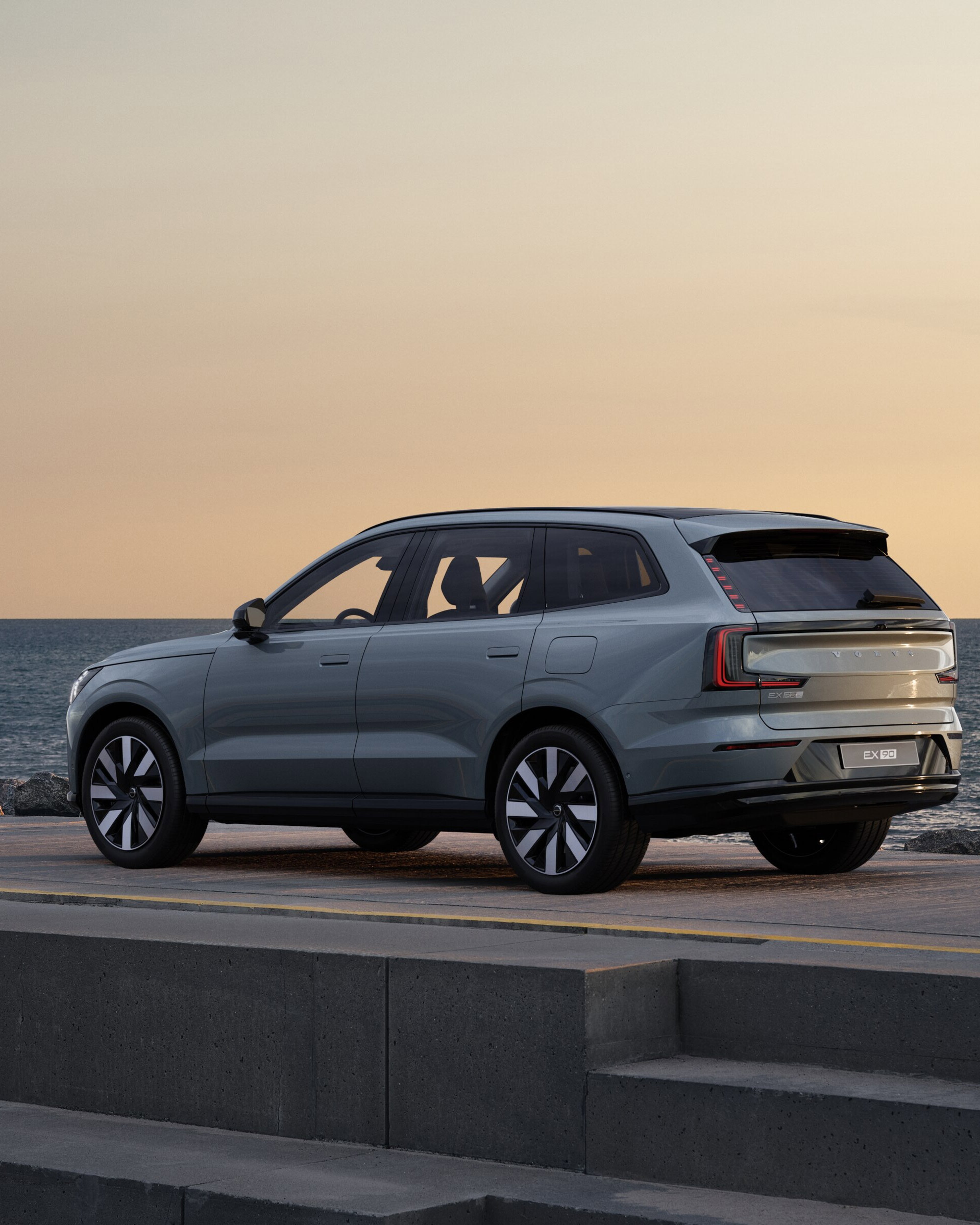 Vue latérale d'un Volvo EX90 gris à une station de charge sur fond orange pastel.