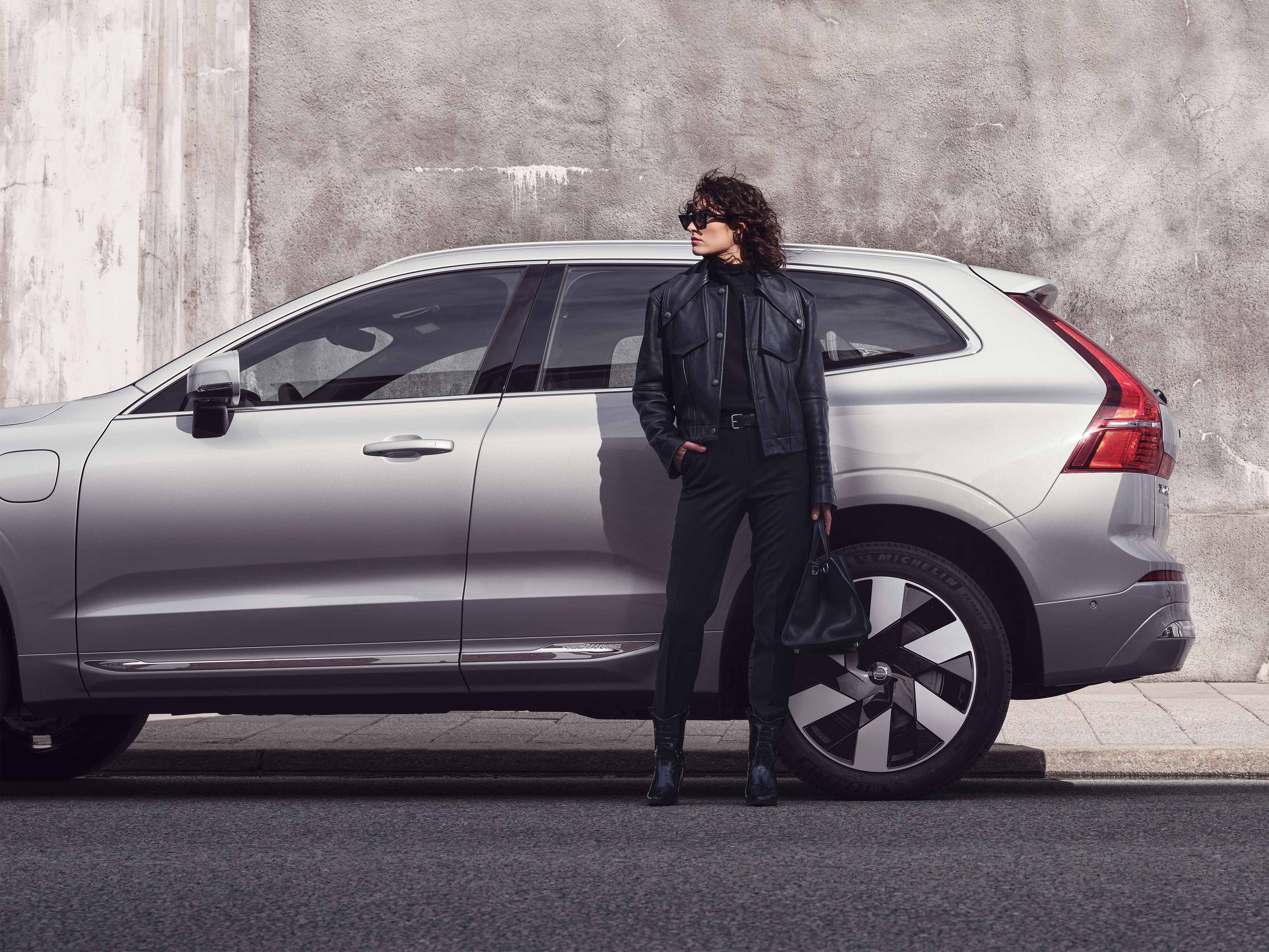 Aurinkolaseihin ja nahkatakkiin pukeutunut nainen seisoo pysäköidyn Silver Grey värisen Volvo XC60 auton takaoven edessä.