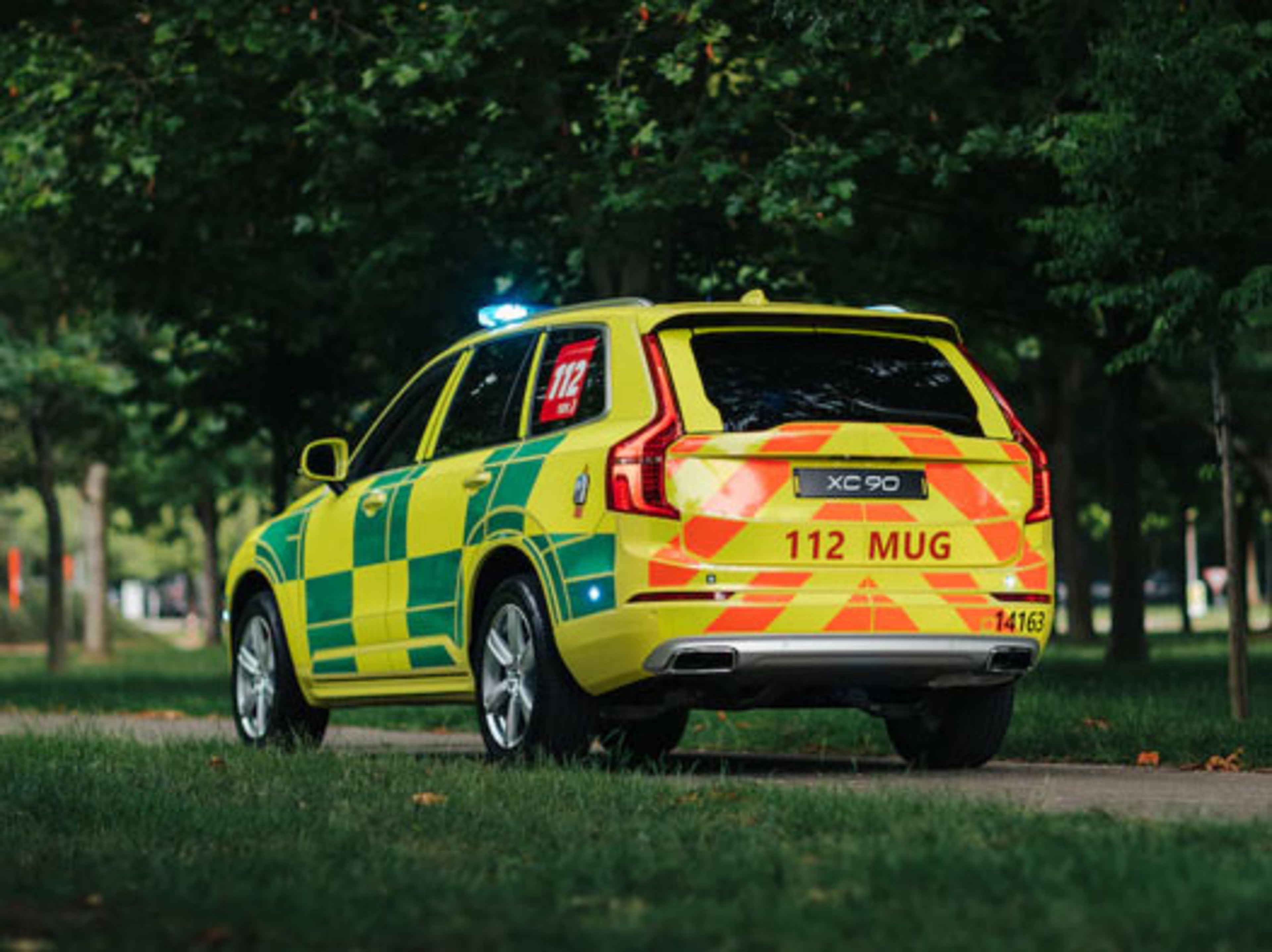 Volvo XC90 d'urgence jaune et verte, plaque 112 MUG, stationnée sur un chemin dans un parc.