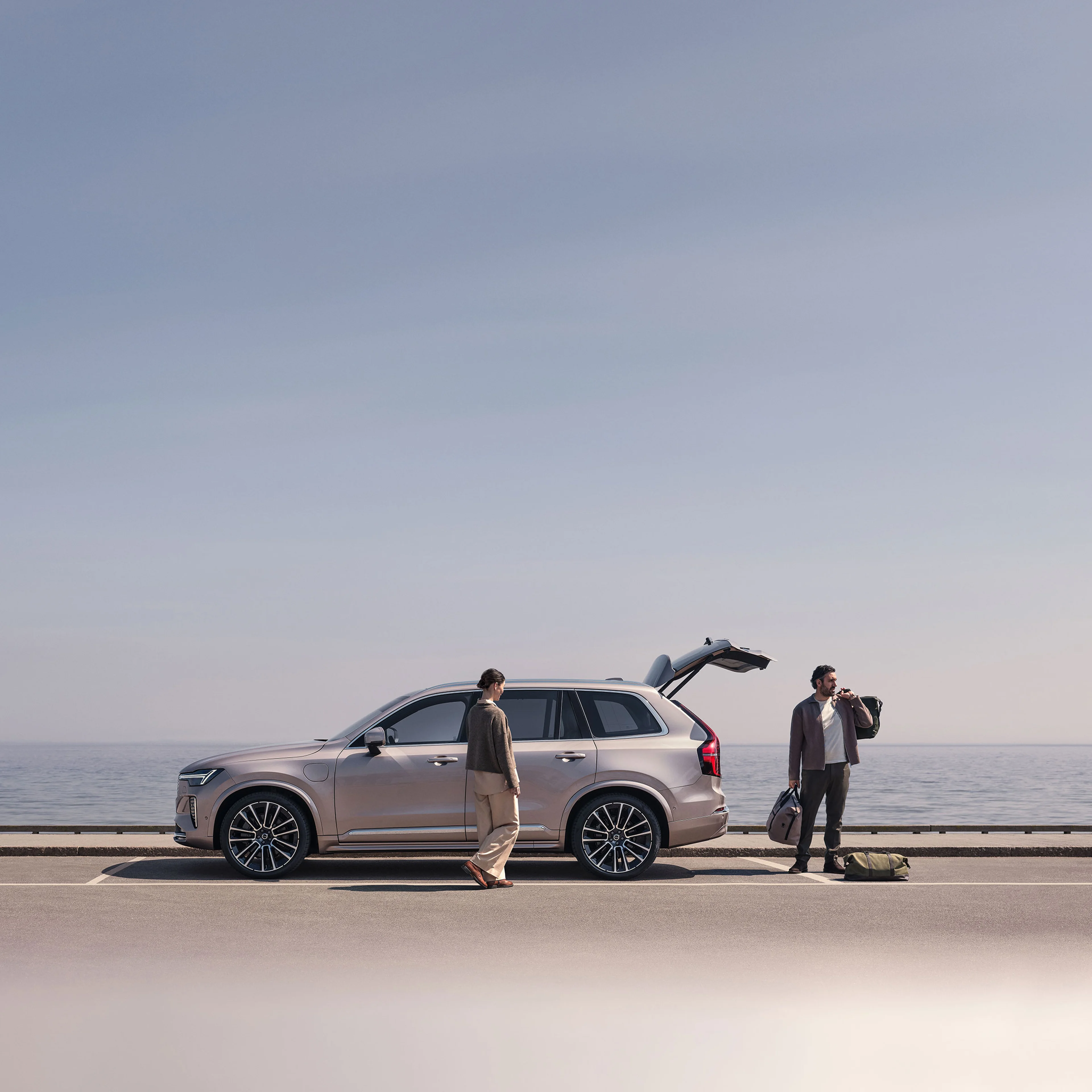 Zwei Personen stehen an einem hellen Volvo XC90 in der Farbe Sand Dune. Der Kofferraum ist geöffnet und die Personen haben Taschen zum einladen dabei.