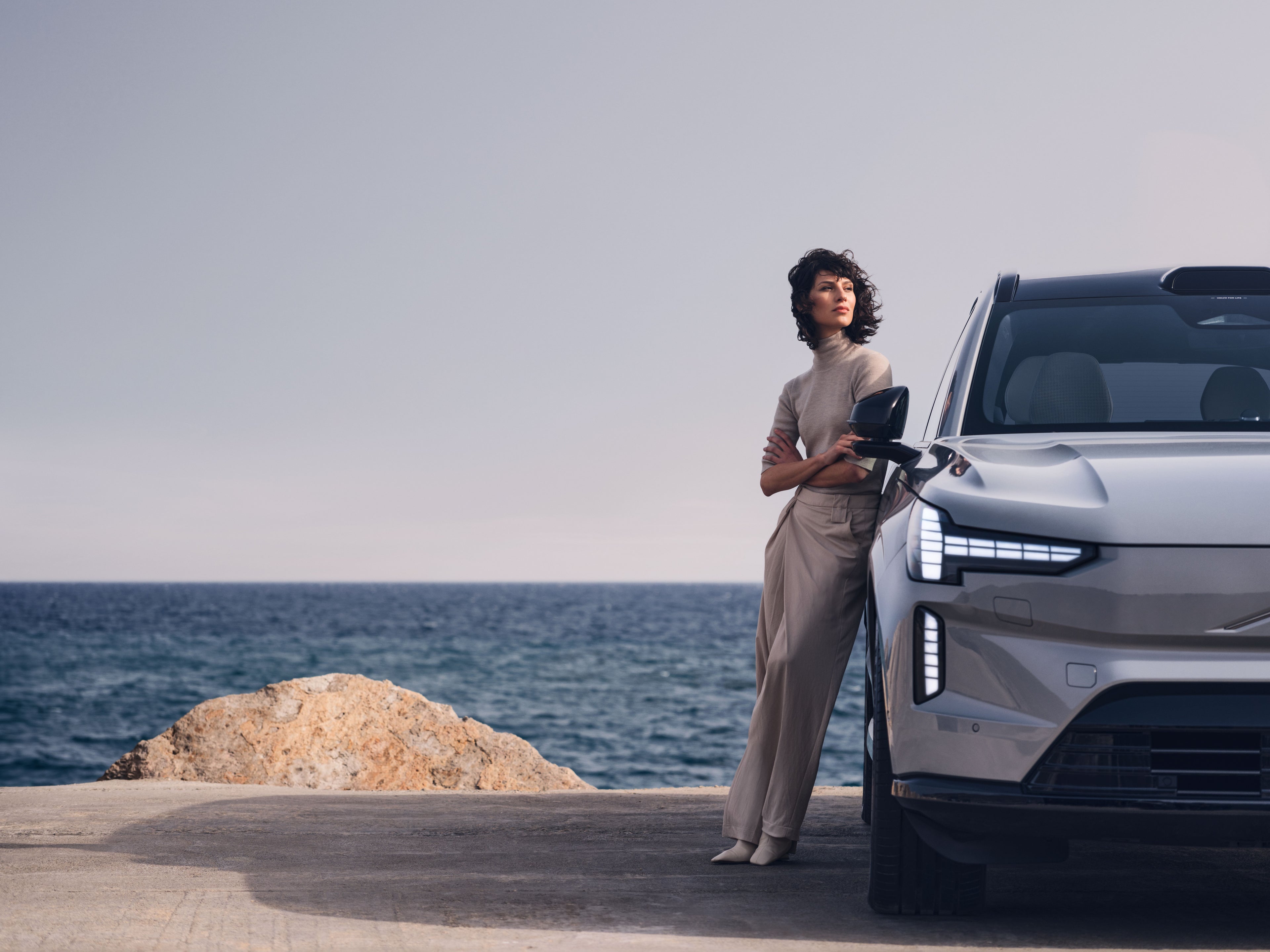 Women standing to the side of our Volvo EX90 in vapour grey with the see behind.