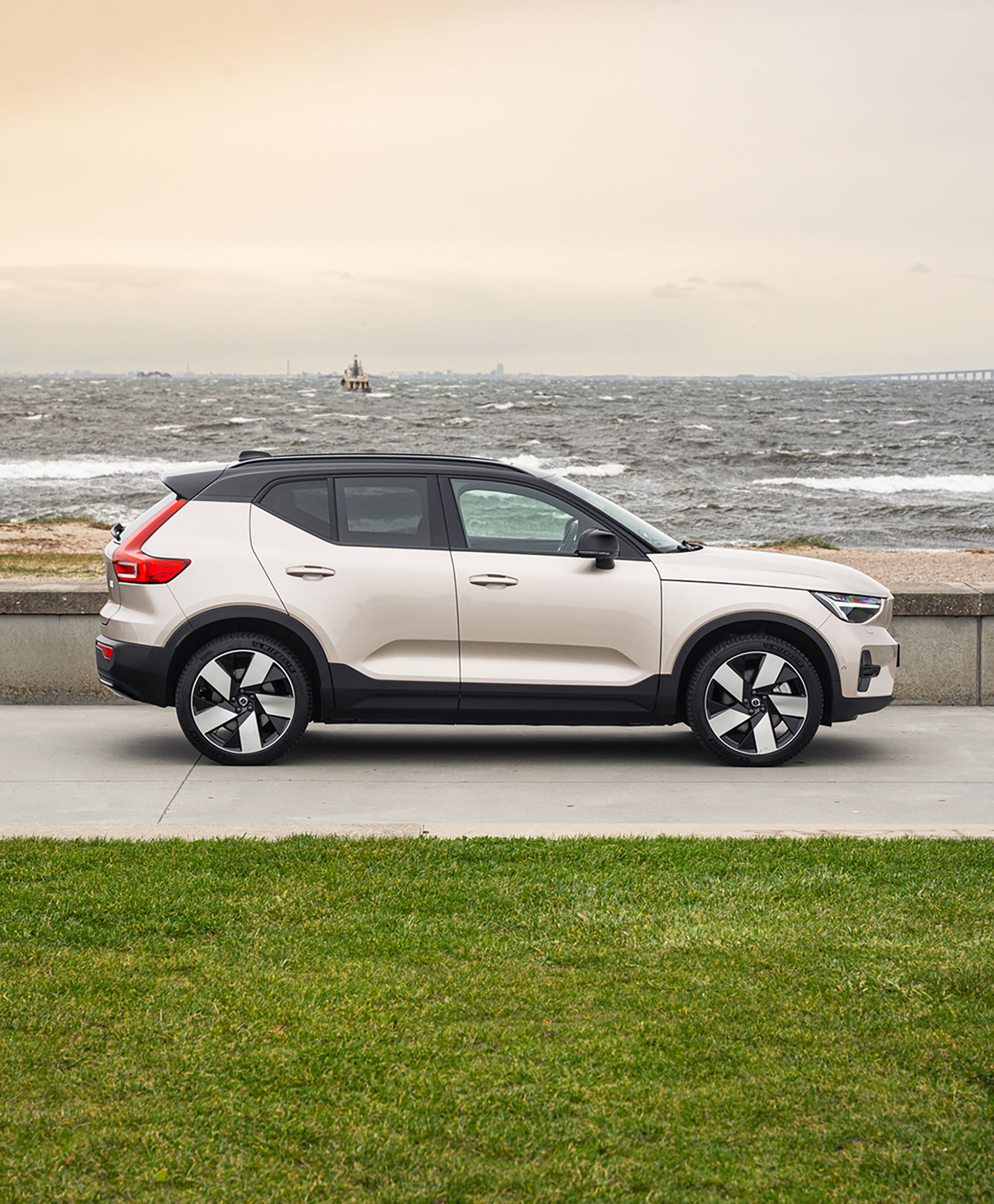 En beige og sort Volvo SUV holder parkeret på en kystvej med oprørt hav og overskyet himmel i baggrunden, hvilket skaber en stemning af eventyr og robusthed.
