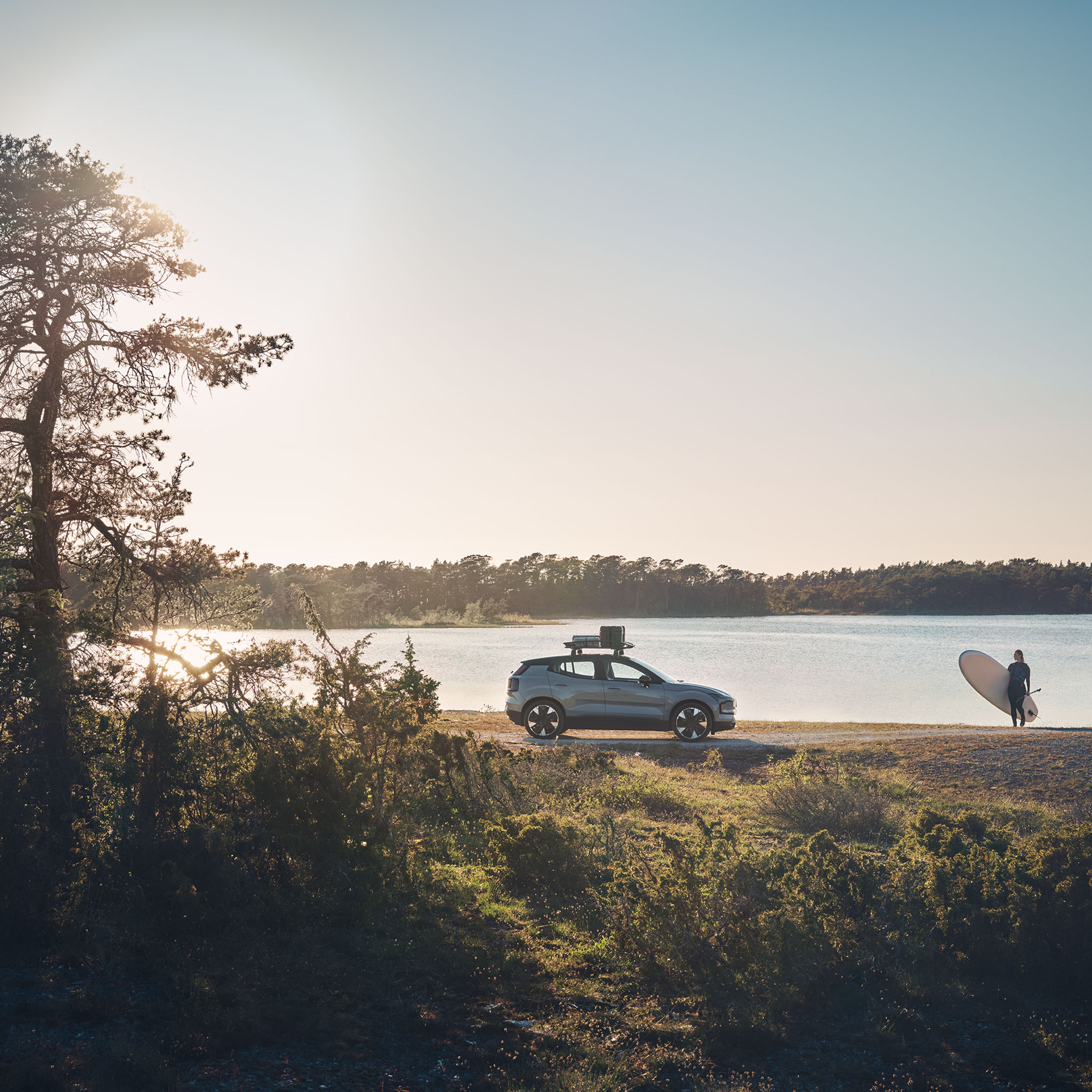 Järven rantaan pysäköity Vapor Grey -värinen Volvo EX30 täyssähköauto ja aurinkoisella rannalla seisova nainen surffilauta kainalossa.