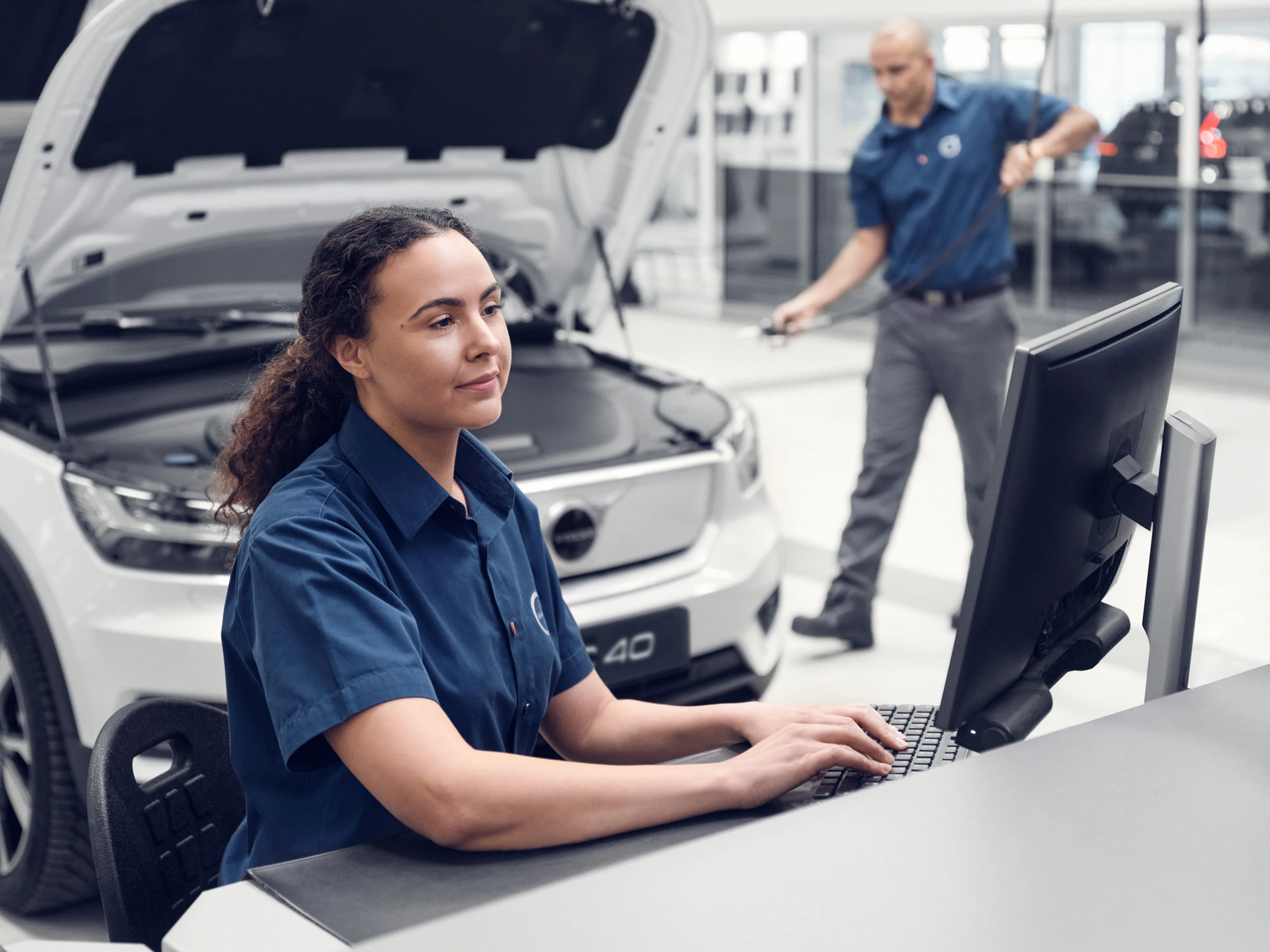 A woman technician works at a computer in a showroom booking in services, while a man inspects a Volvo in the background.