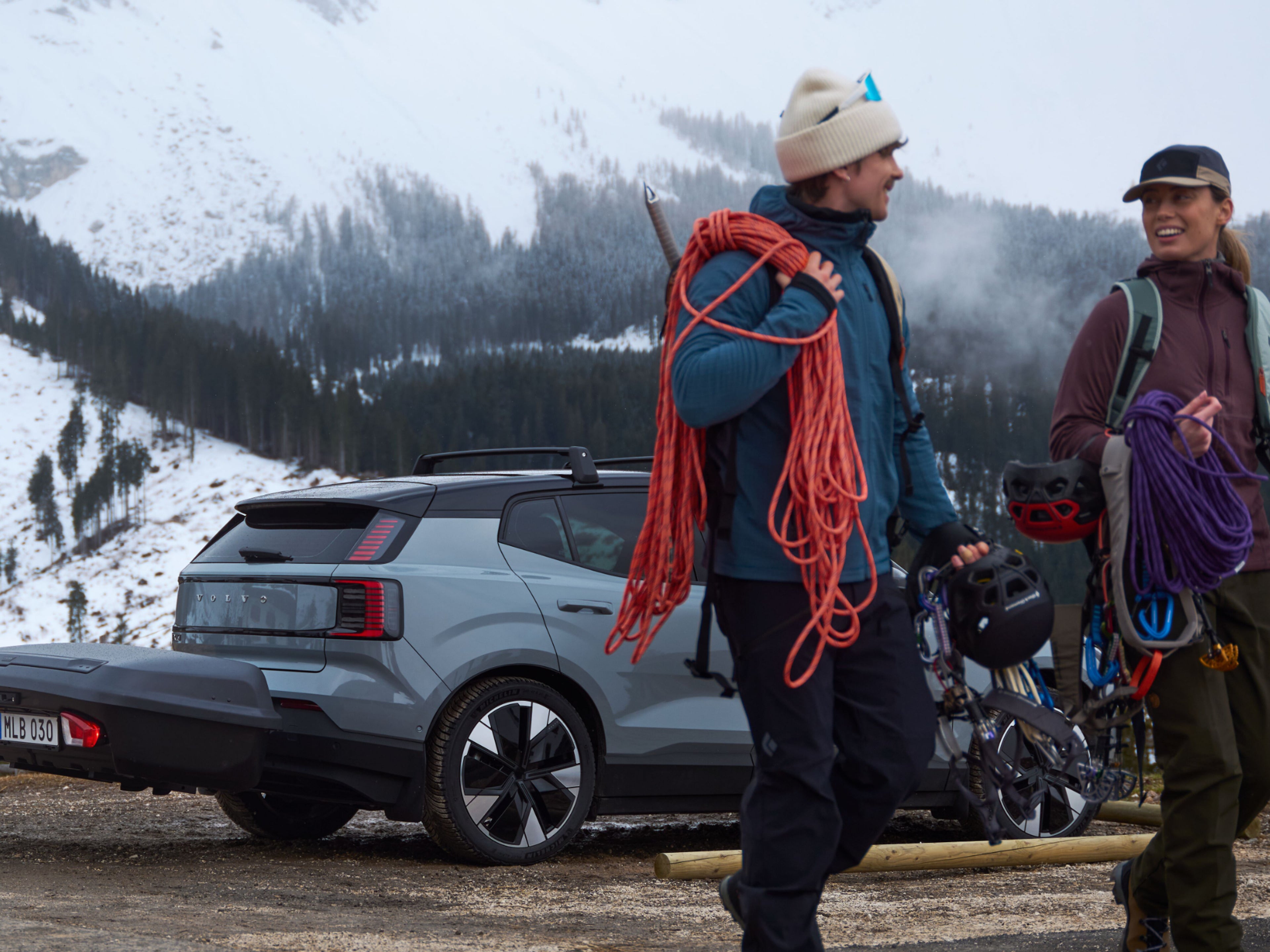 Ein Mann und eine Frau tragen Winterausrüstung und entfernen sich von einem grauen Volvo EX30.