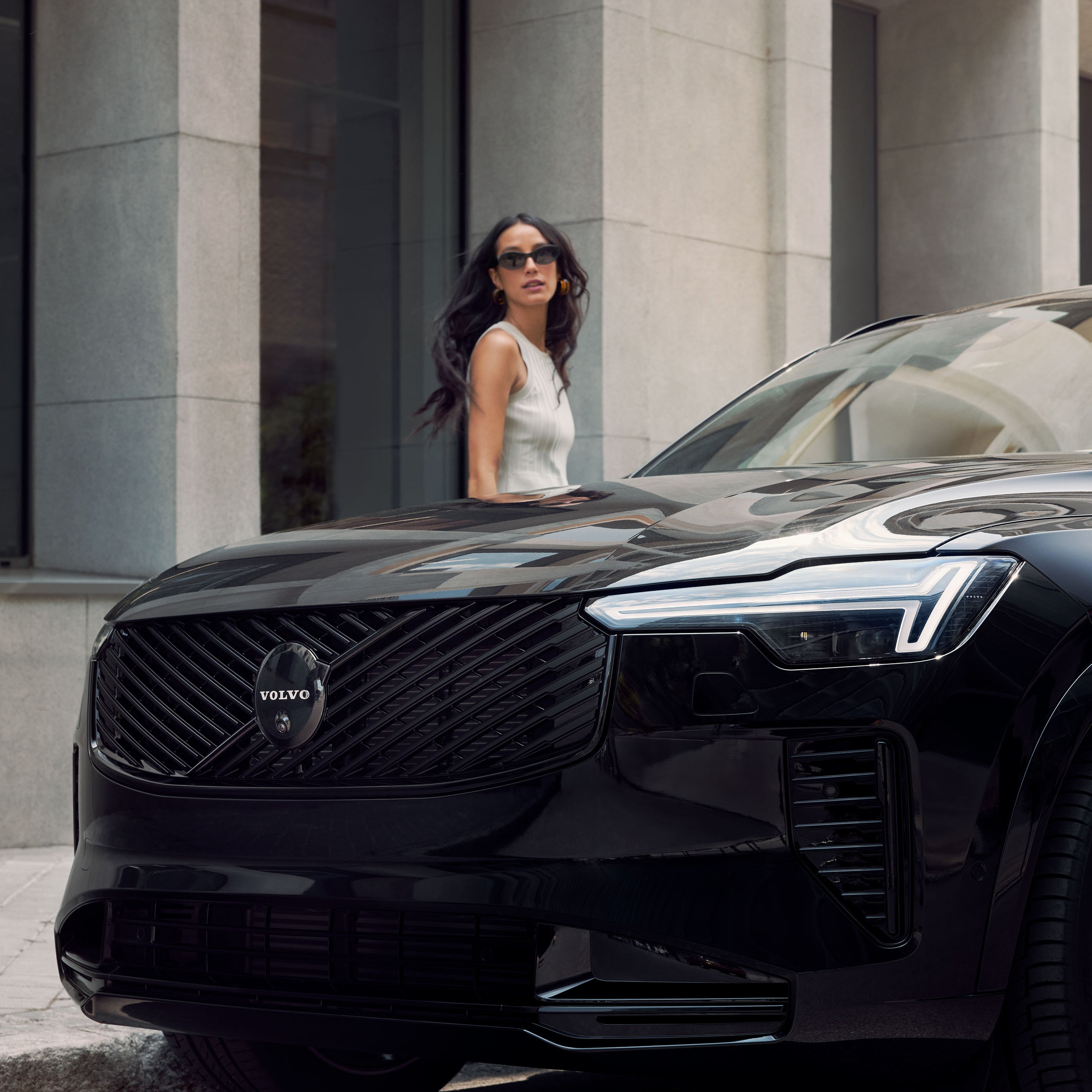 Tummatukkainen nainen seisoo Volvo XC90 Black Edition lataushybridikatumaasturin takana ja katsoo olan yli kohti kameraa.