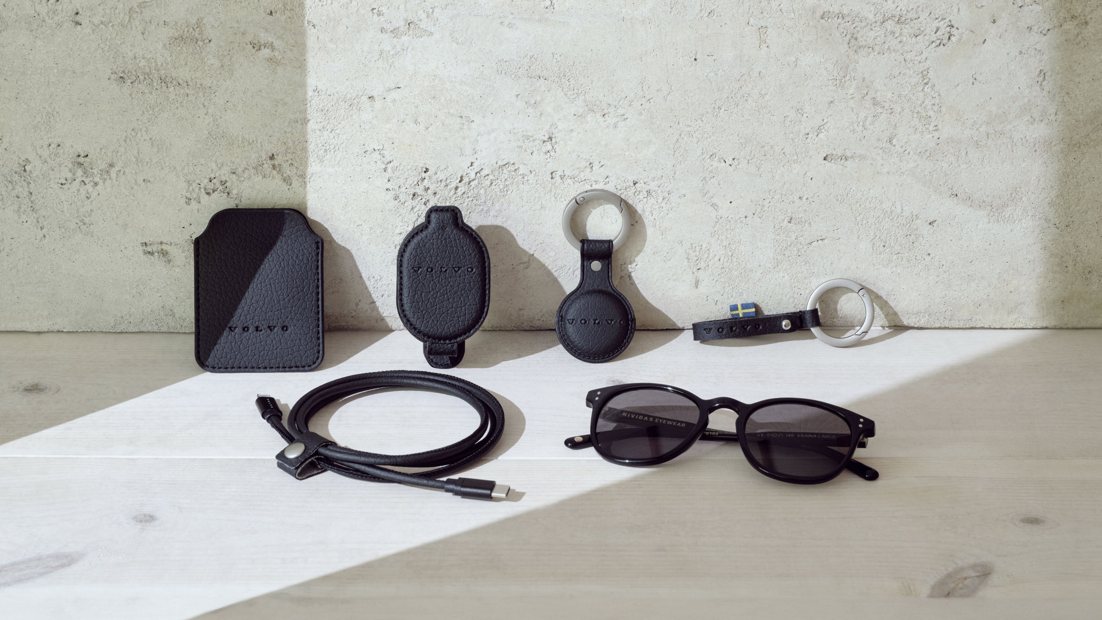 Schwarz mattes Volvo Zuebhör steht aufgestellt vor einer beigen Wand. Darunter Ladekabel, Volvo Sonnenbrille, Key Tag, Hüllen für Handy und Schlüssel