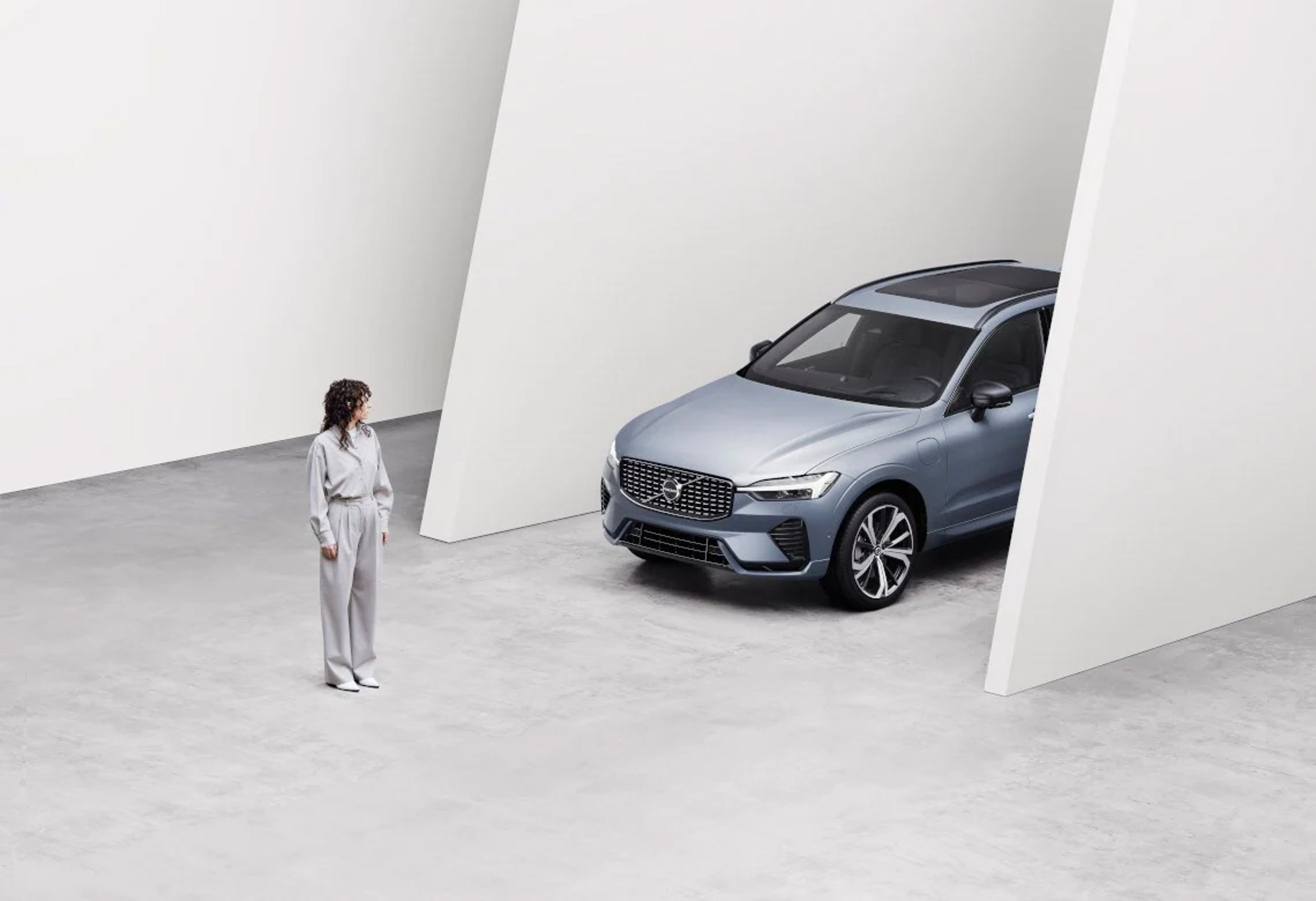 Eine Frau steht vor einem blau-grauen Volvo XC60 SUV in einem großen, weißen Raum.