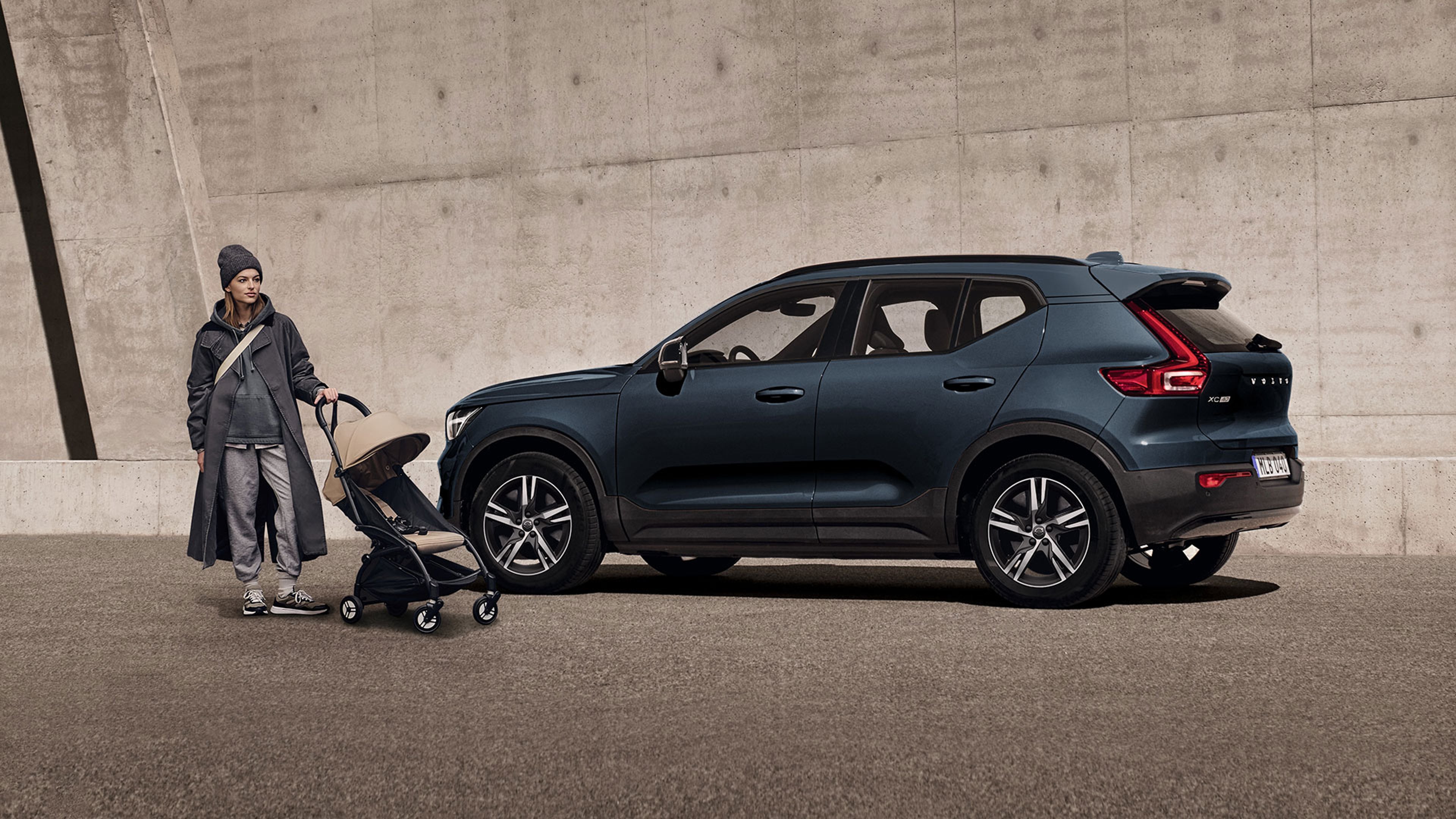 Une femme en imperméable tenant une poussette se tient debout devant un XC40 Volvo bleu