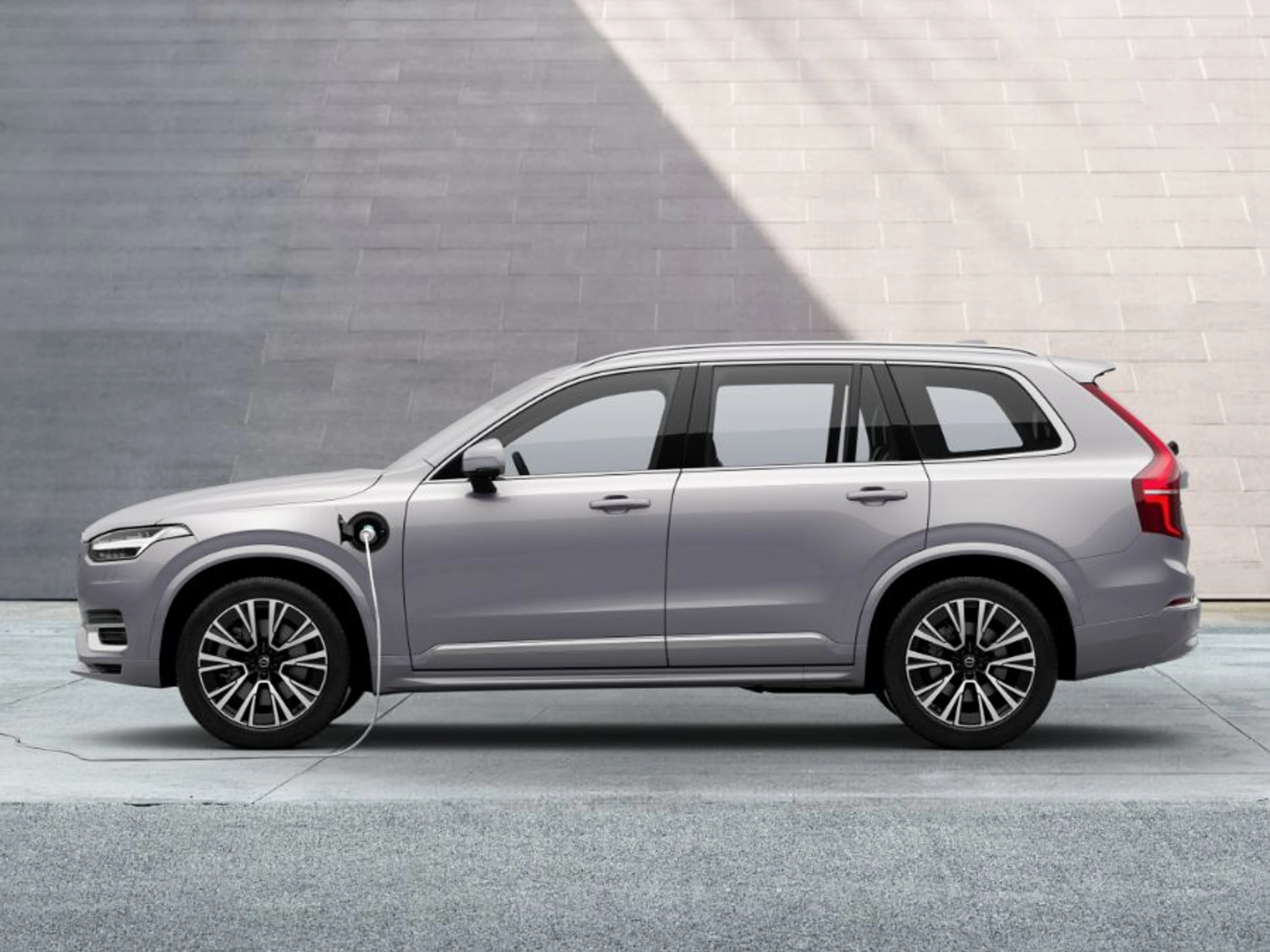 Crni Volvo XC90 Plug-in Hybrid SUV, priključen na punjenje s vidljivim kablom, parkiran u prostranom krajoliku pod blagim svjetlom, simbolizirajući njegovu snažnu prisutnost i ekološku odgovornost.
