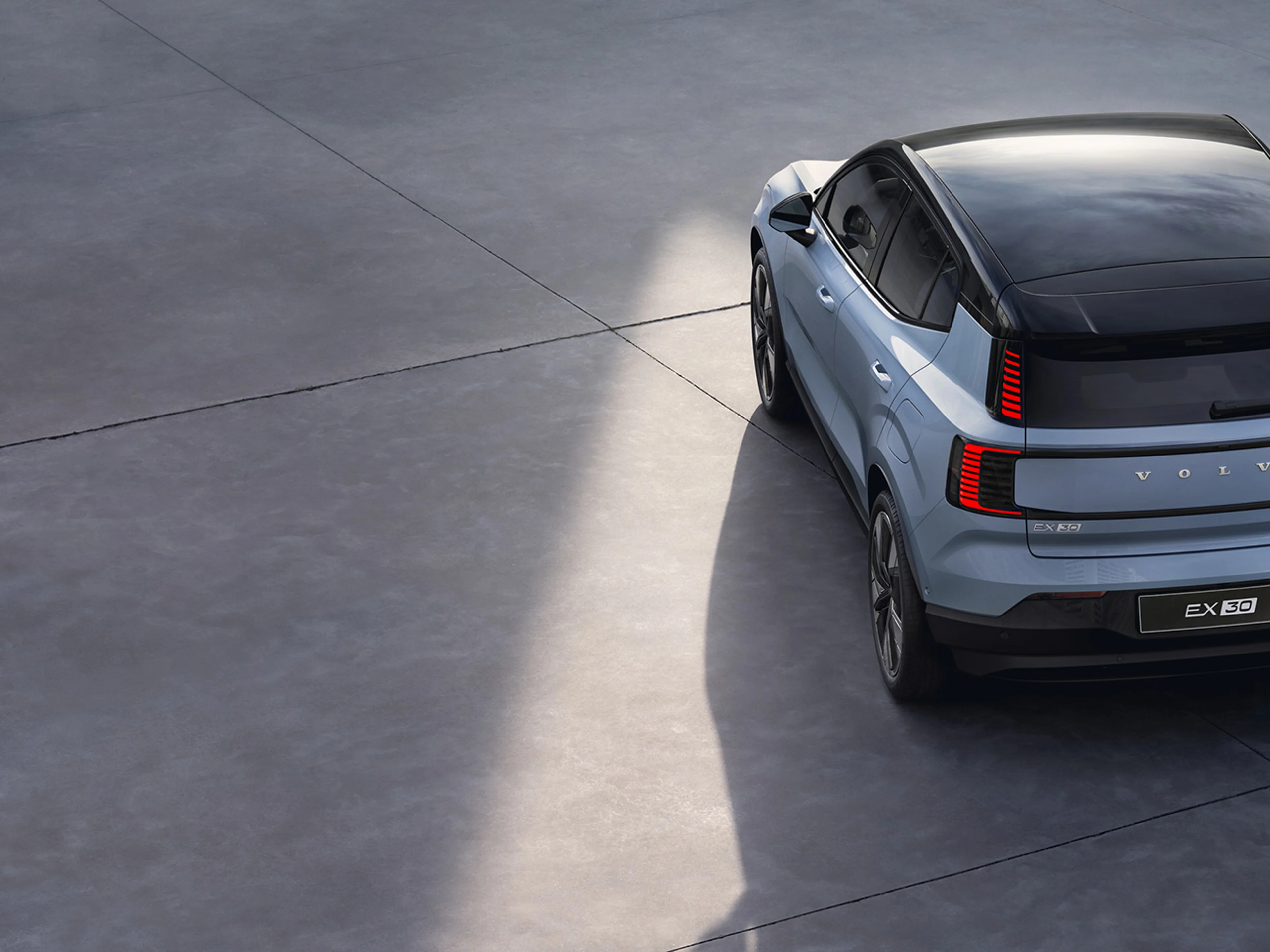 A top view of a cloud blue Volvo EX30 electric SUV parked on a smooth, light grey surface, showcasing its sleek design and black roof.