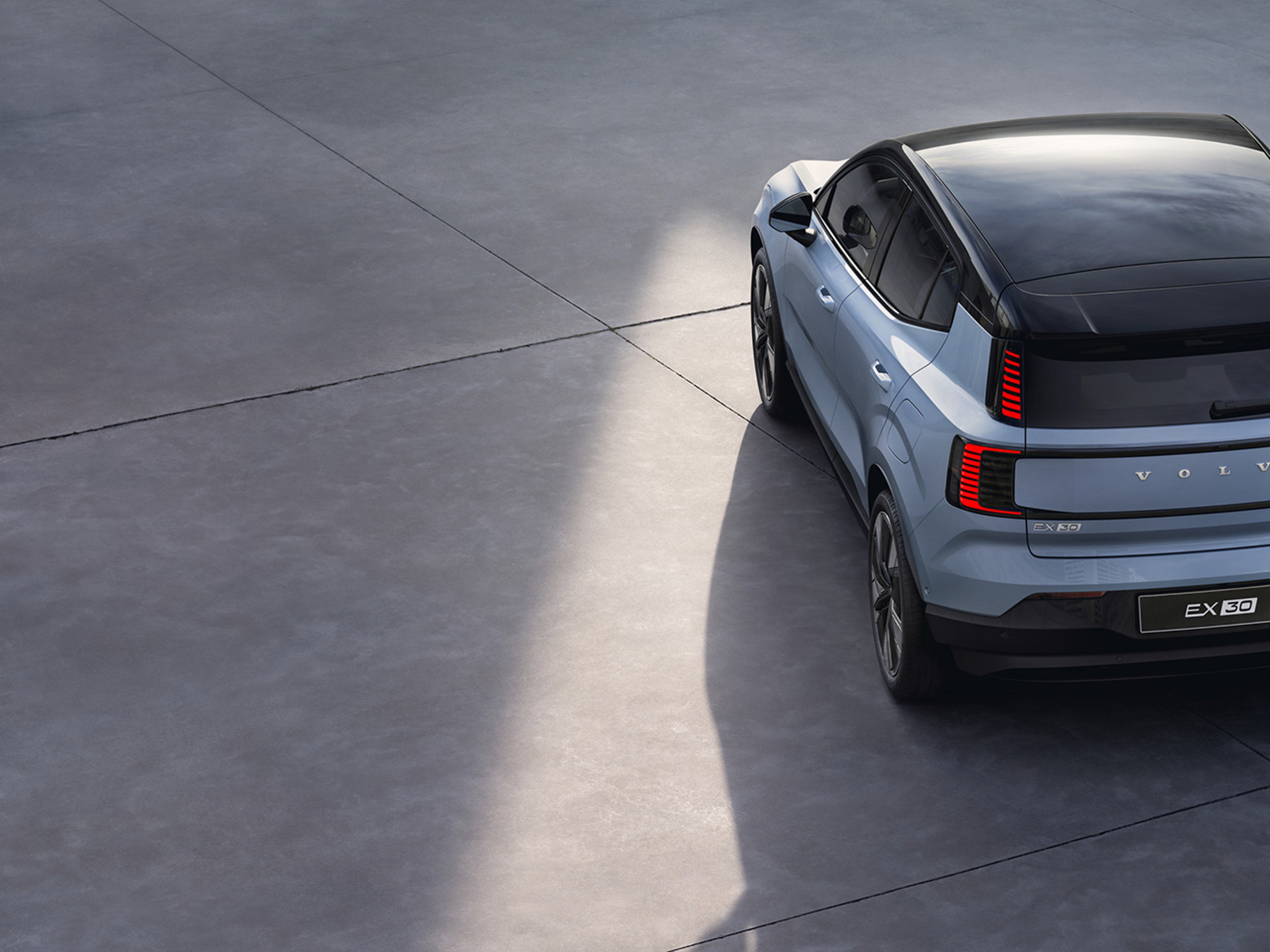A top view of a cloud blue Volvo EX30 electric SUV parked on a smooth, light grey surface, showcasing its sleek design and black roof.