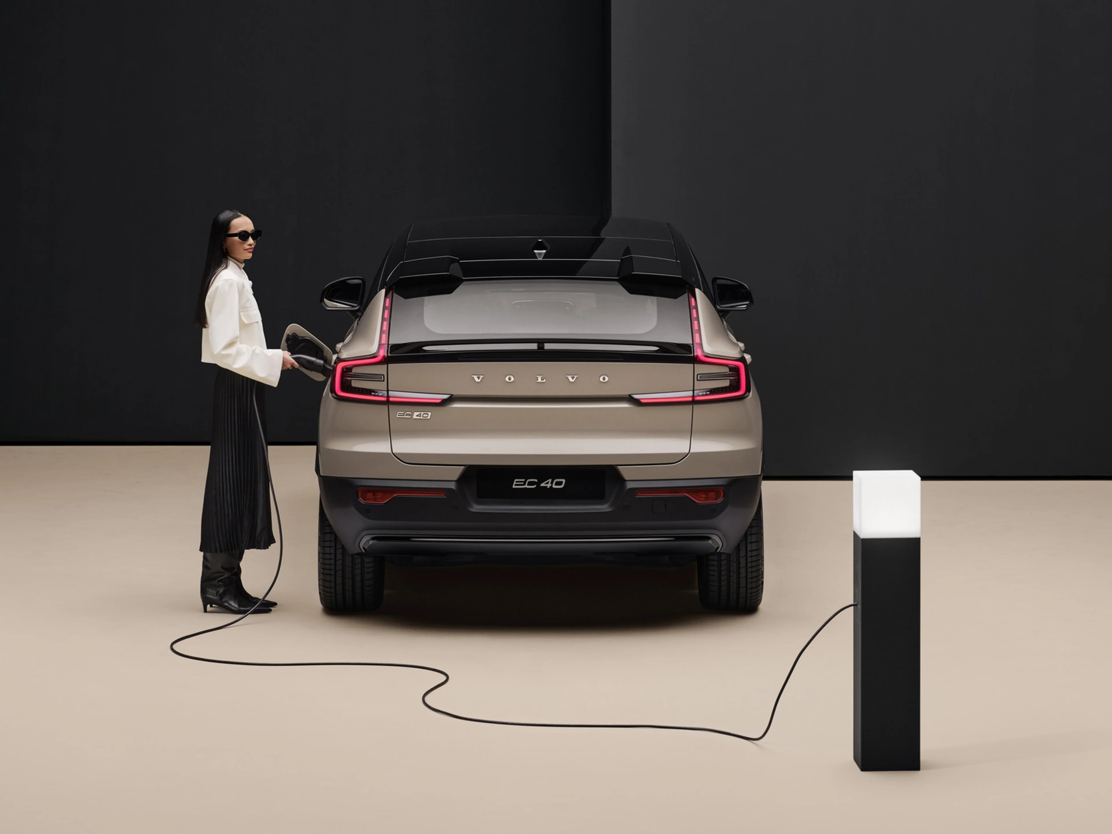 A rear profile of the fully electric Volvo EC40 in colour Sand Dune , with a woman plugging it in to charge. There is a beige and black backdrop.