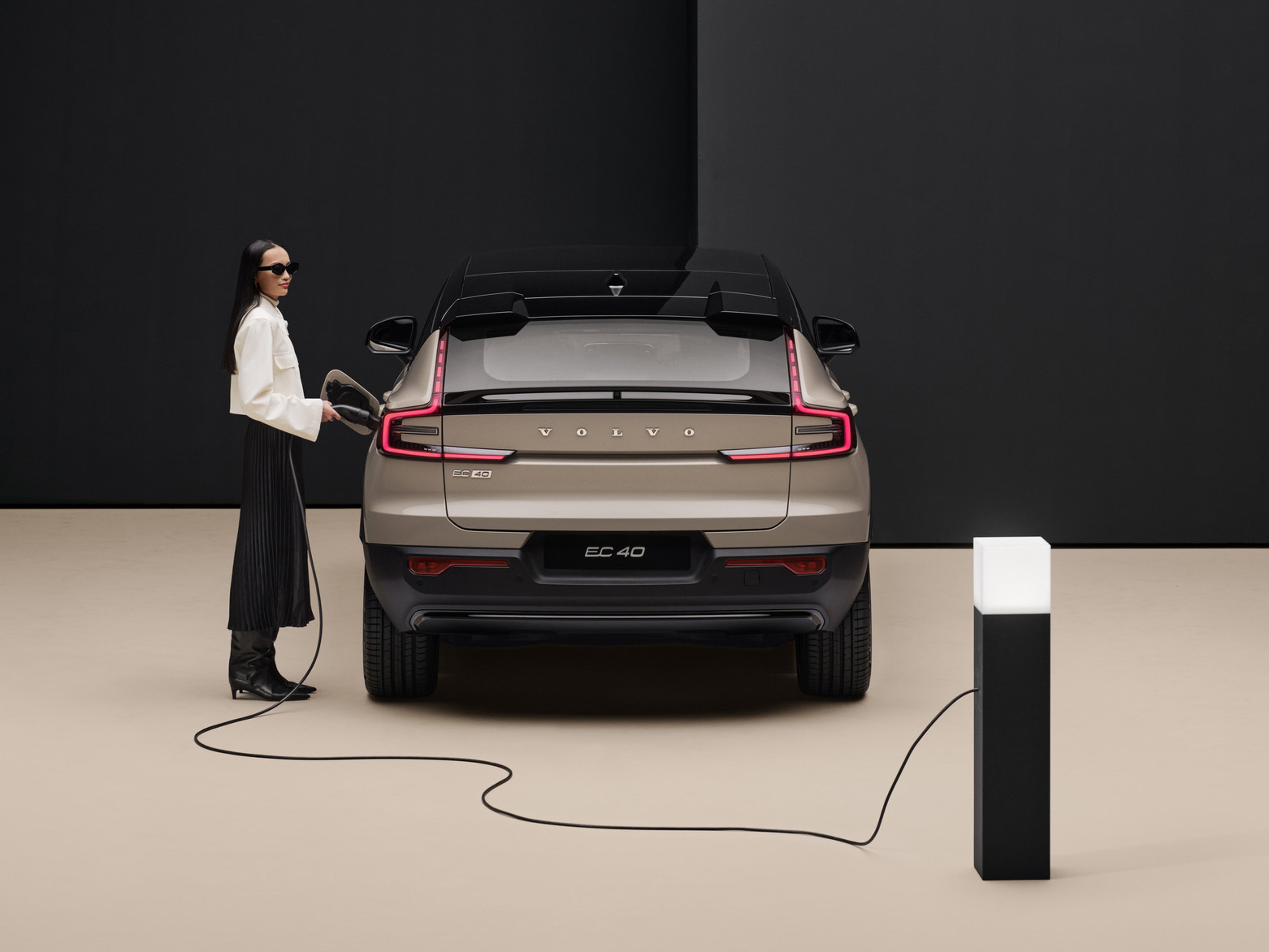 A rear profile of the fully electric Volvo EC40 in colour Sand Dune , with a woman plugging it in to charge. There is a beige and black backdrop.