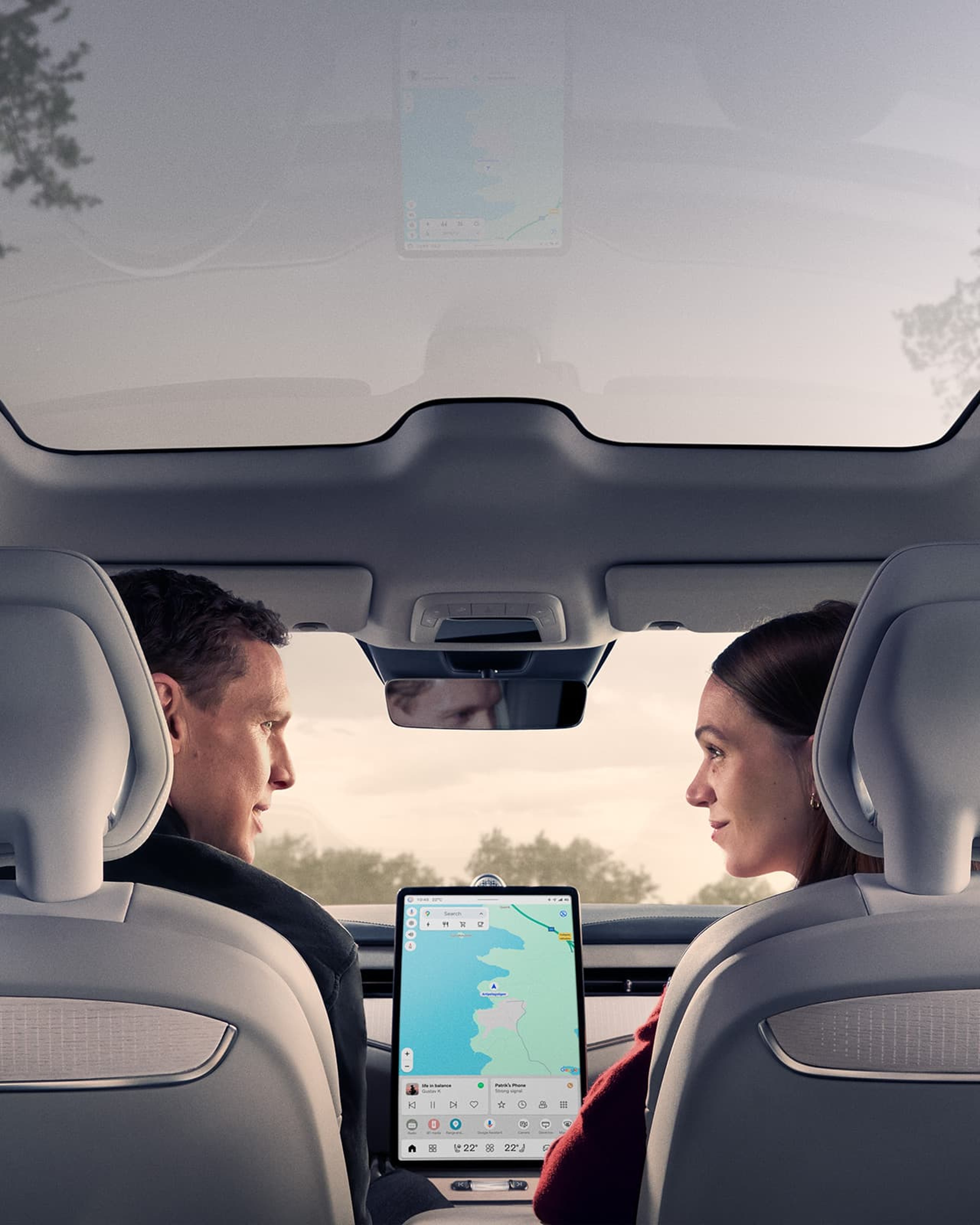 Un homme et une femme sont assis dans une Volvo ES90 avec un toit panoramique et regardent la tablette sur le tableau de bord.