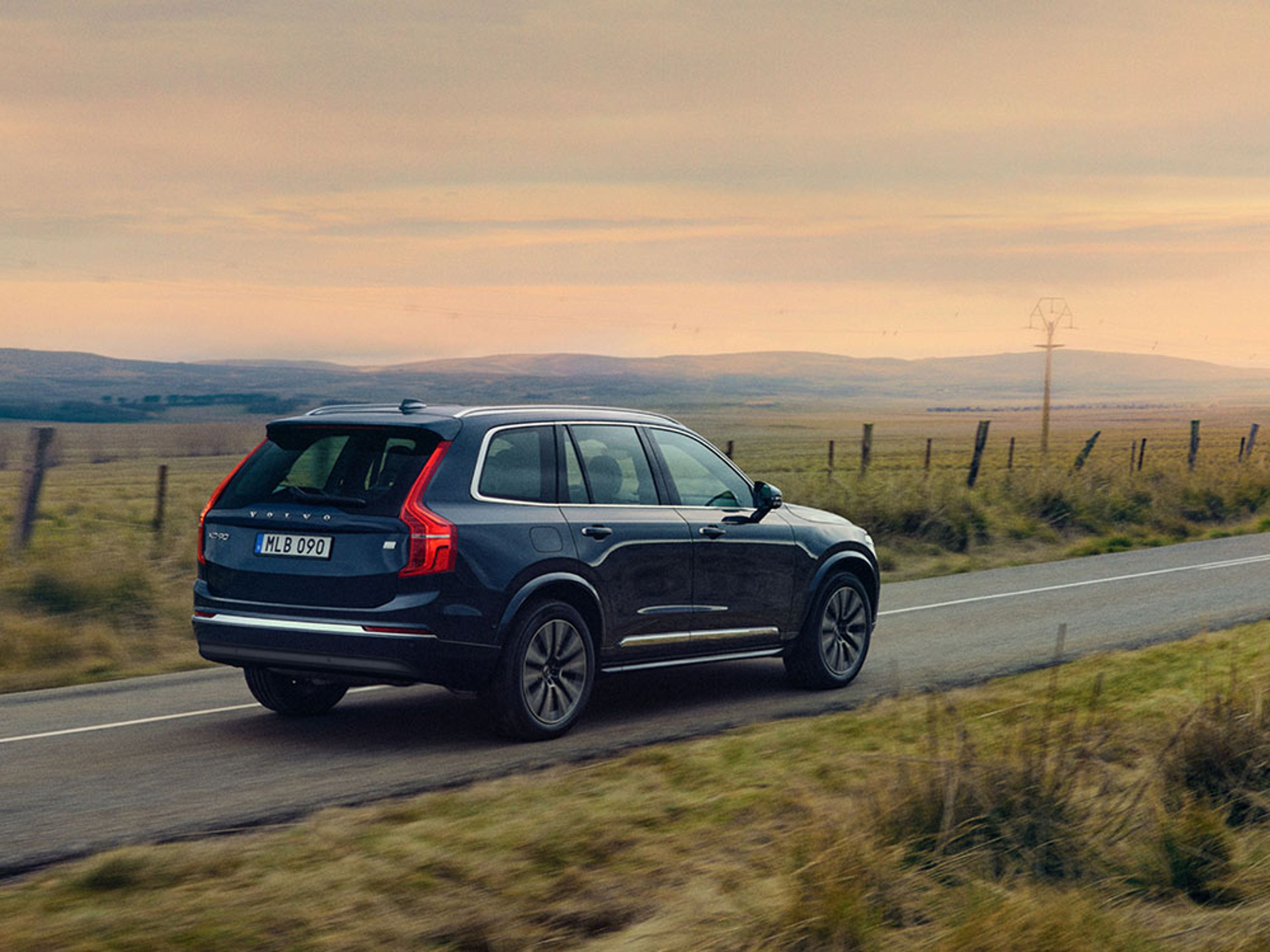 Een donkerblauwe Volvo XC90 rijdend op een landweg met heuvels en een zonsondergang op de achtergrond.