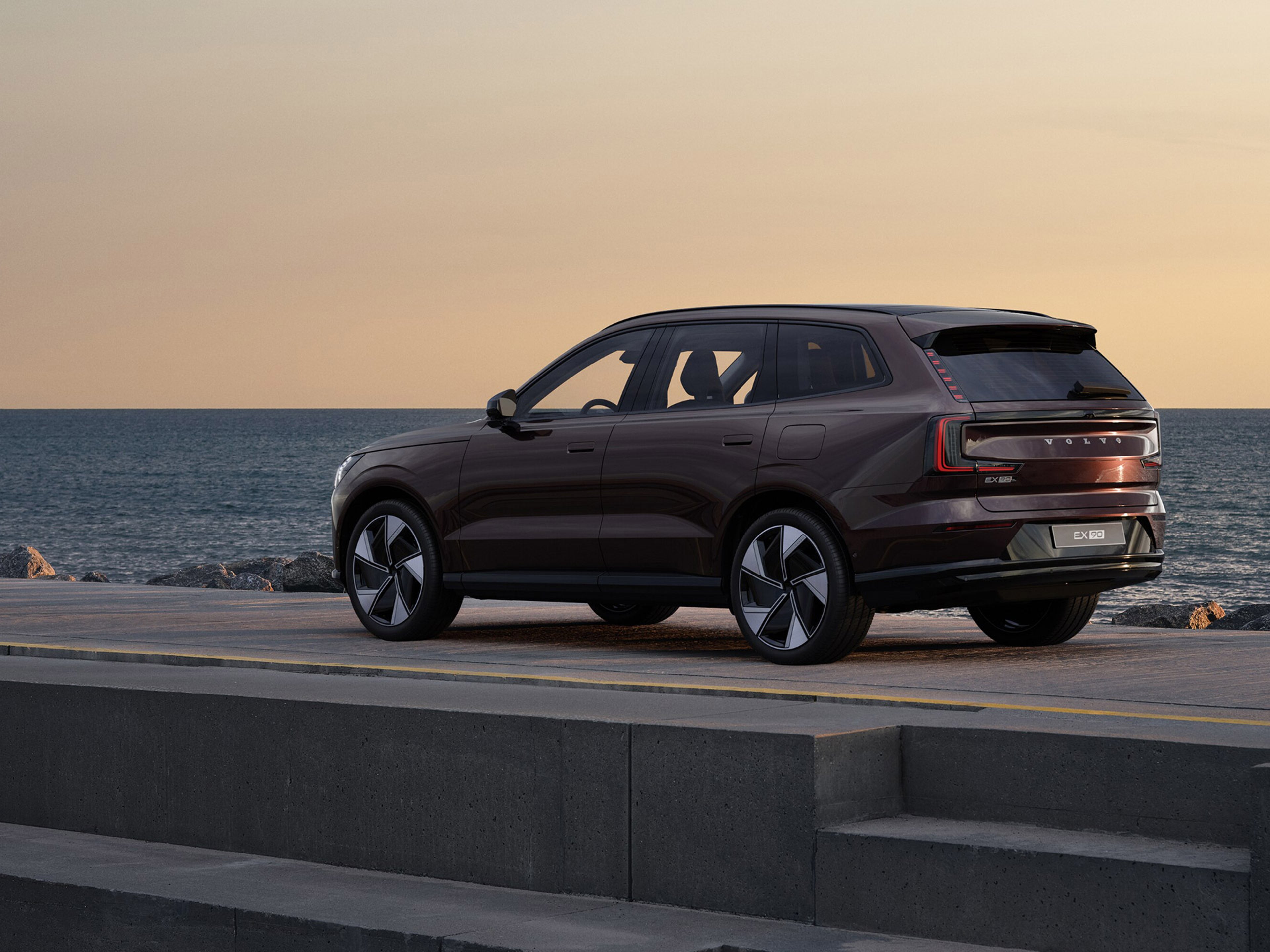 Mulberry red -värinen Volvo EX90 täyssähköauto pysäköitynä rantaan katukiveykselle auringonlaskussa.