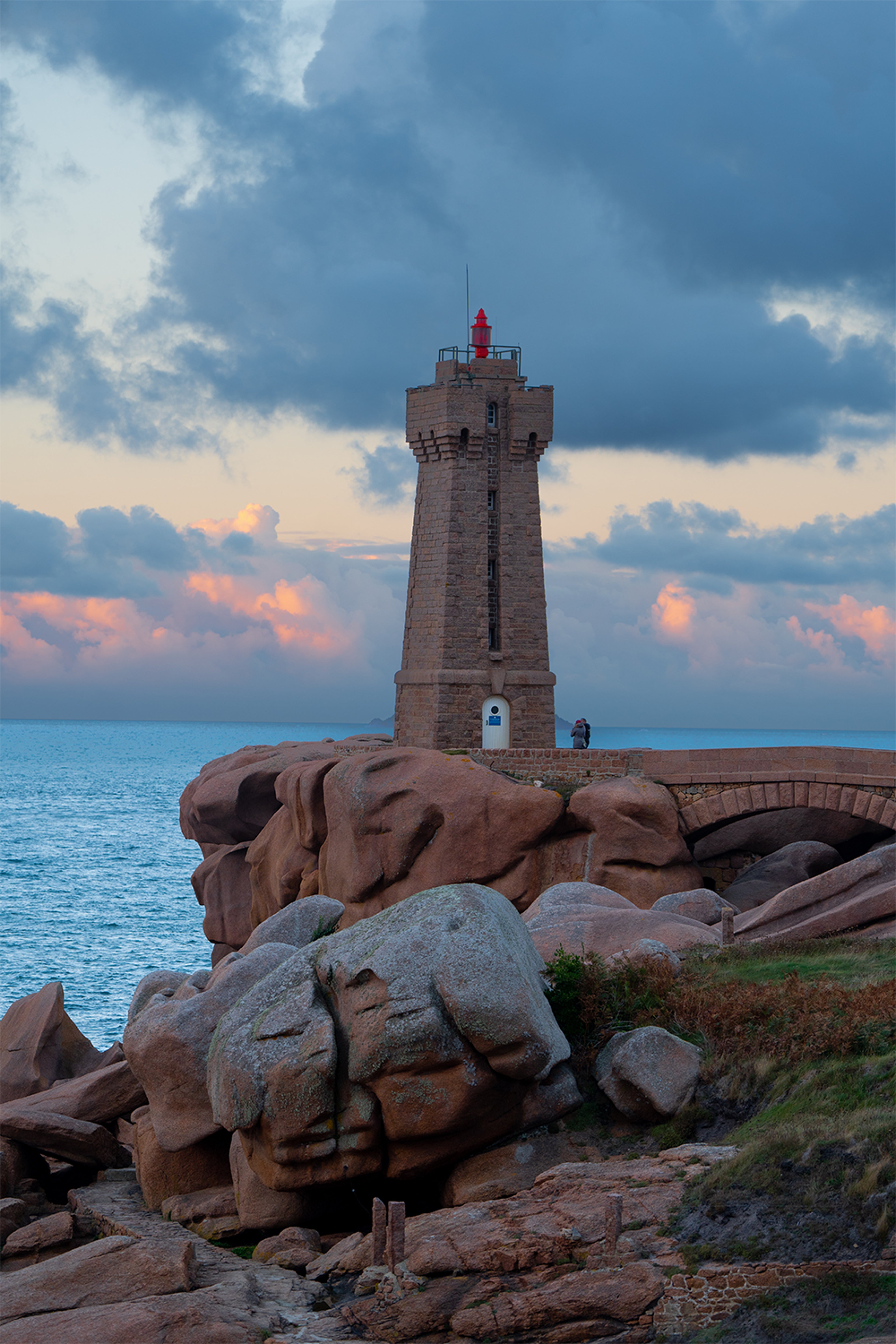 Phare en granit rose sur côte rocheuse au coucher du soleil, ciel nuageux et mer en arrière-plan.