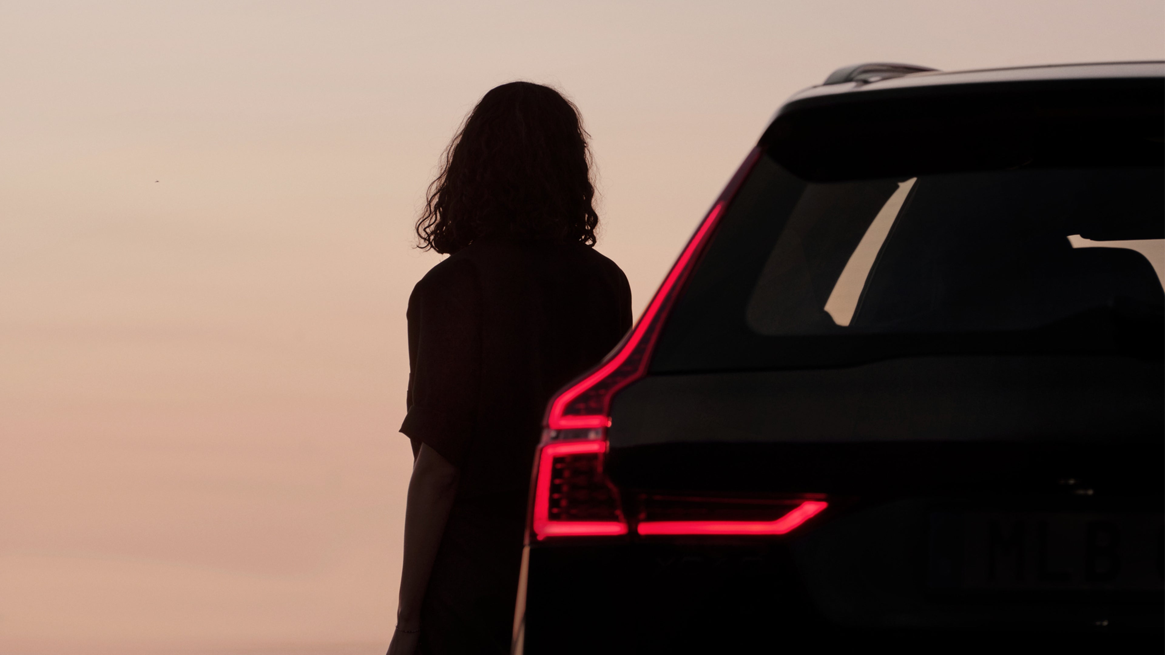 Naisen siluetti ja Volvo XC60 Black Edition lataushybridin vasen takakulma auringonlaskussa.
