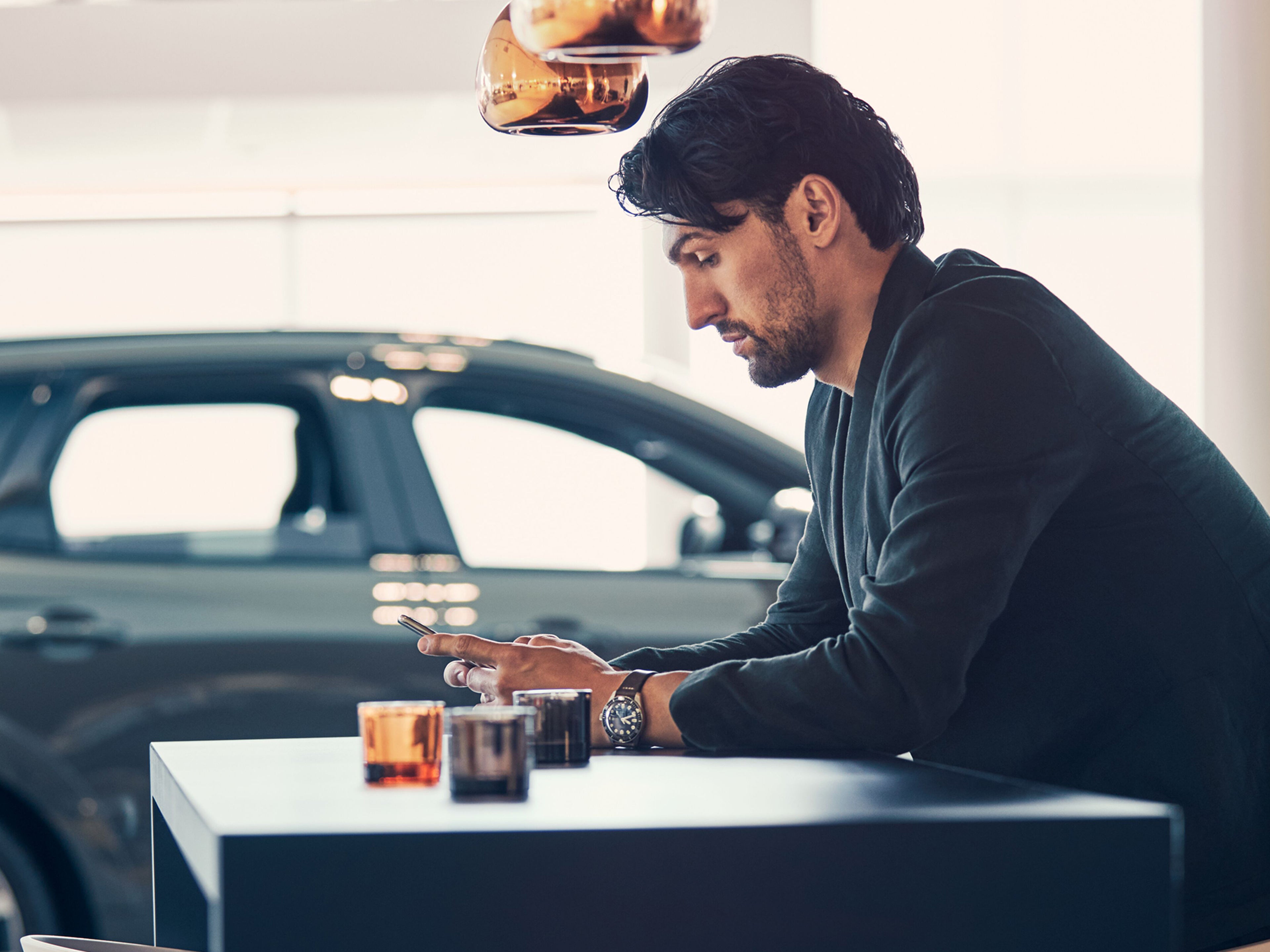 homem sentado no concessionário volvo a verificar o seu telemóvel com um volvo em sendo plano