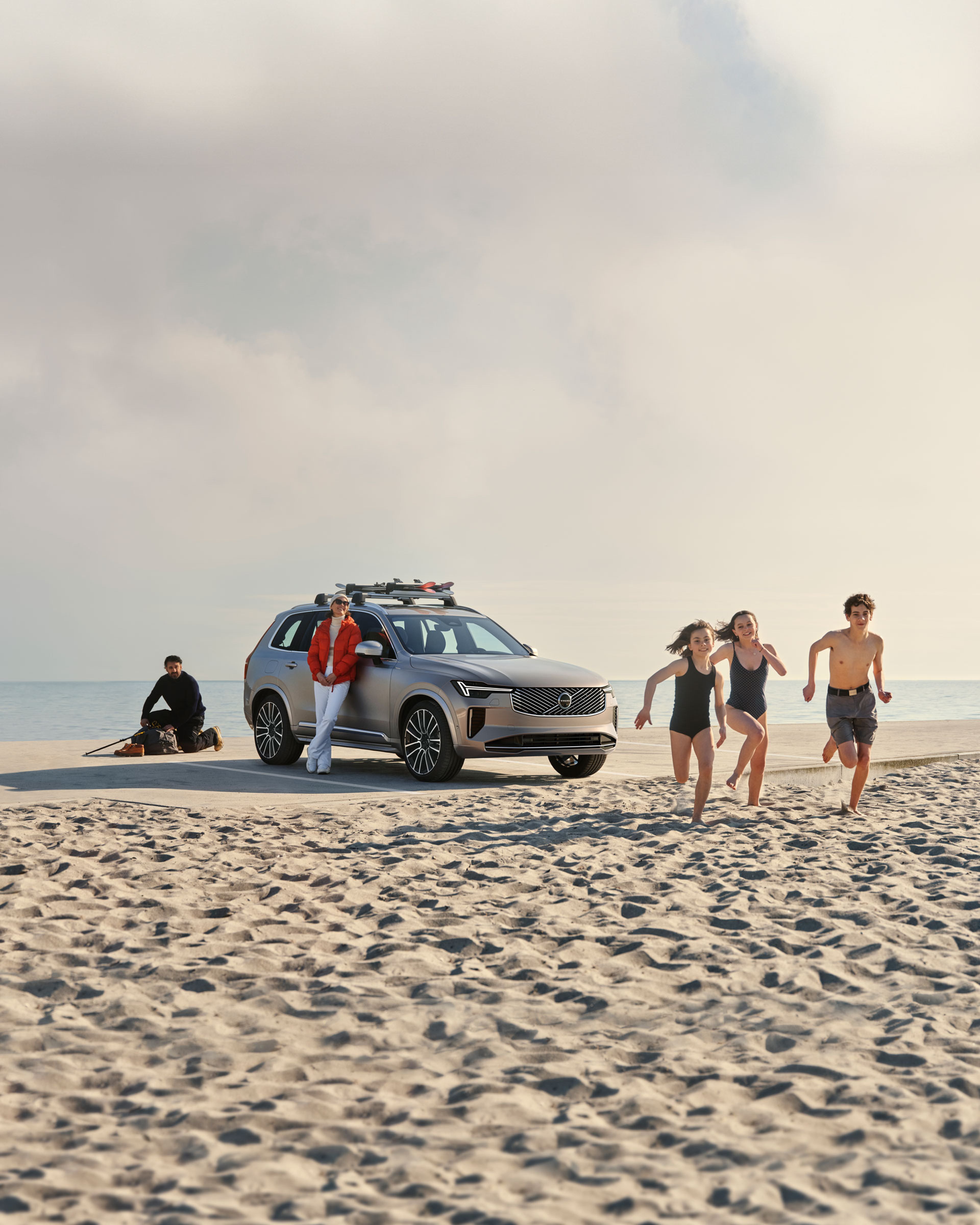 Voiture Volvo XC90 sur une plage, personne appuyée contre elle, trois enfants courant sur le sable.