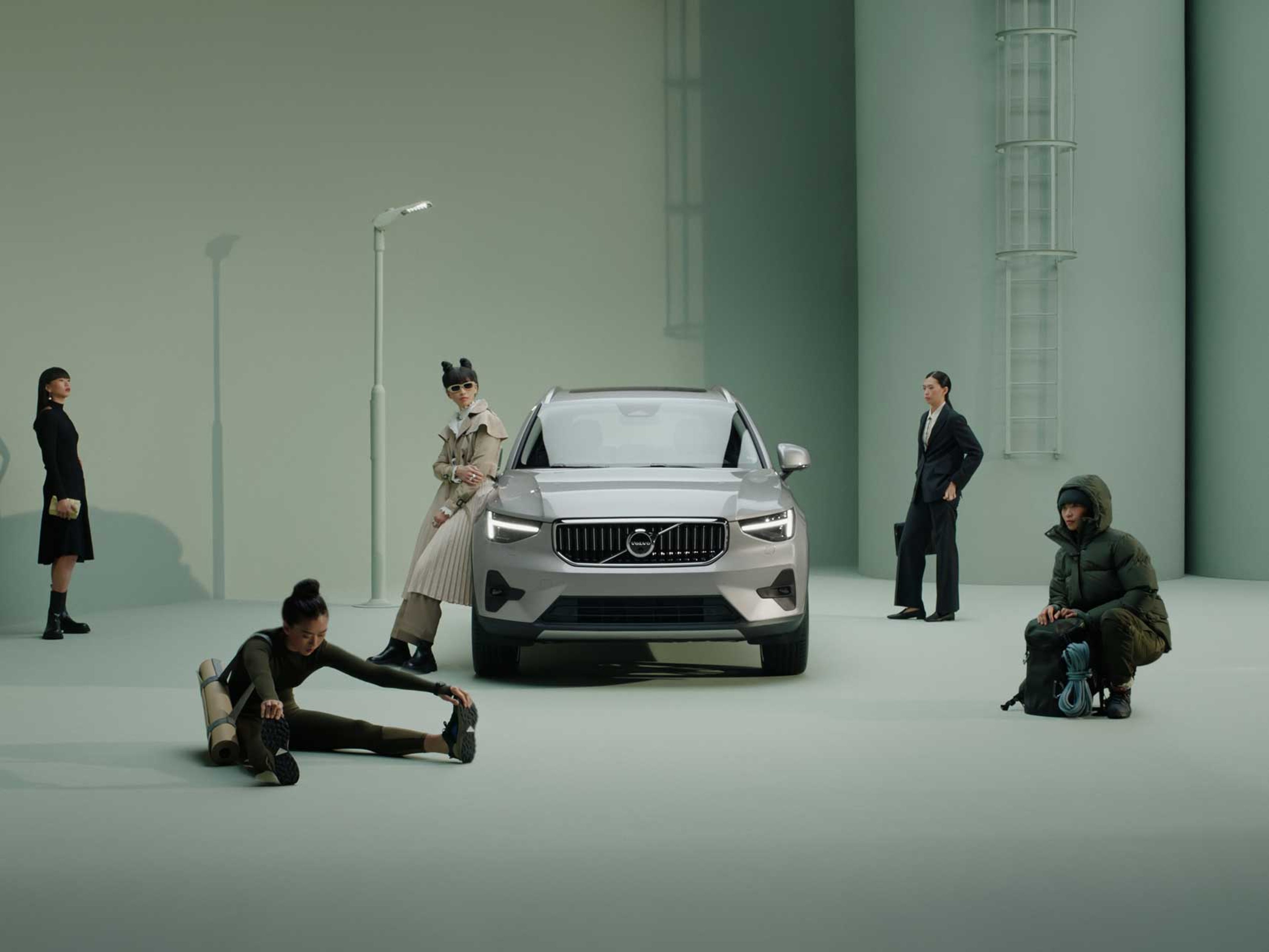 Fem personer i stilfuldt tøj omgiver en elegant sølvfarvet Volvo i en minimalistisk scene. Nogle poserer eller strækker sig og udstråler en smart, moderne æstetik.