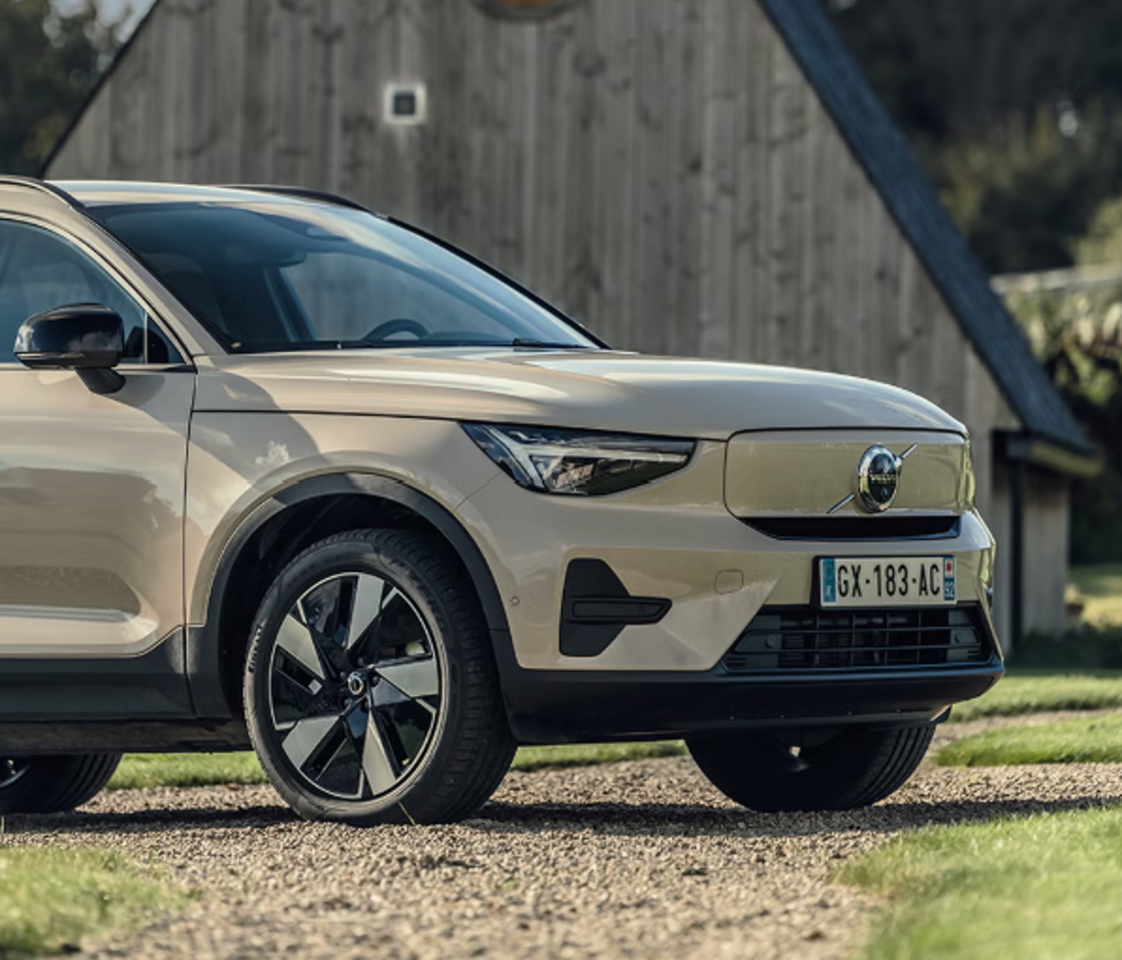 Volvo beige devant maison en bois triangulaire, ciel bleu et nuages blancs en arrière-plan.