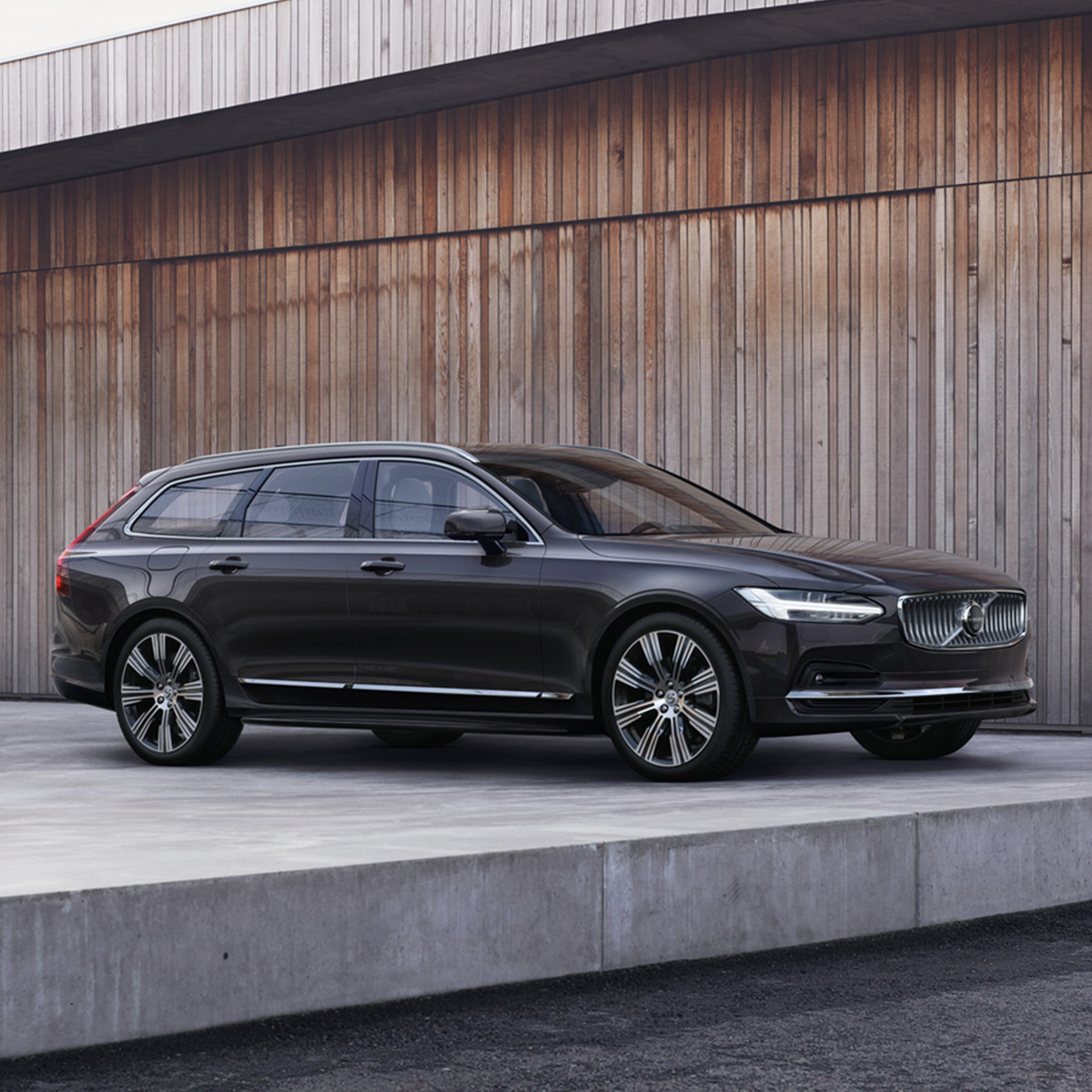 Platinum Grey -värinen Volvo V90 lataushybridifarmari pysäköitynä puisen rakennuksen sisäpihan tasanteelle.