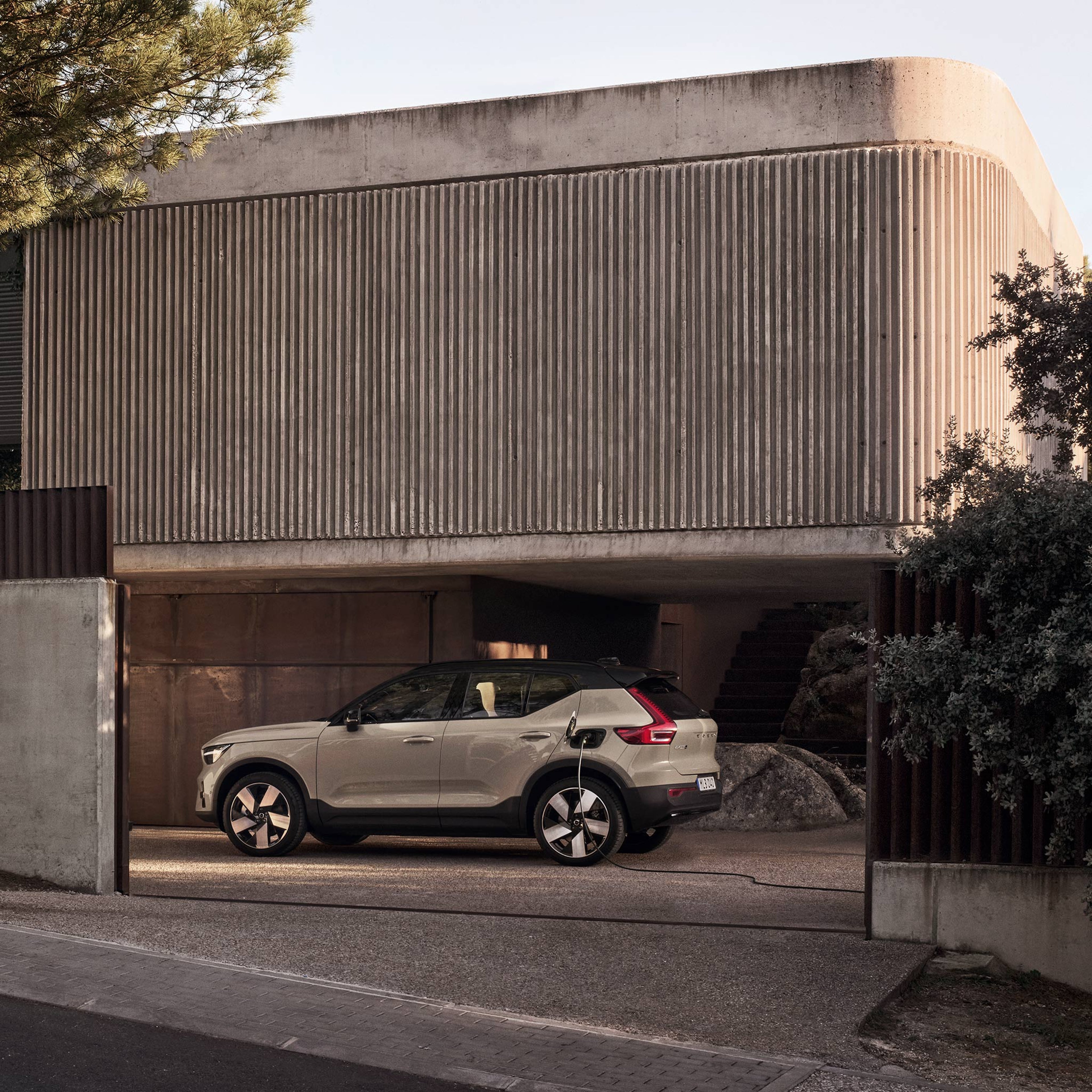 Lataukseen kiinnitetty Sand dune -värinen Volvo EX40 täyssähköinen katumaasturi betonirakennuksen edustalla. Etualalla betoninen aita, jossa kulkuväylä pihalle, ja vehreitä pensaita.