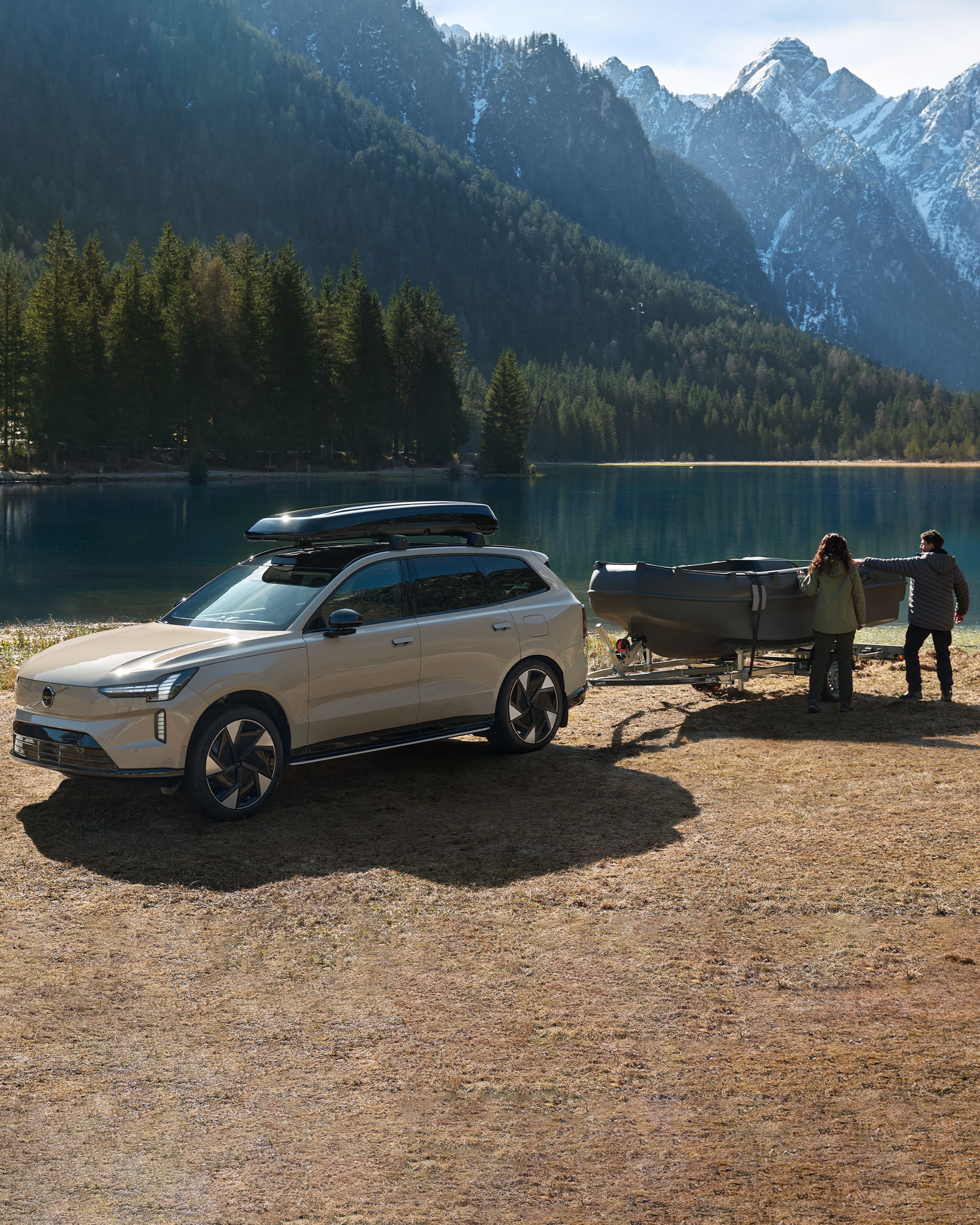 Ein Volvo EX90 in der Farbe Sand Dune steht mit einem Boot Anhänger am Heck angehängt an einem Bergsee zwischen Sandstrand und Bergen. Zwei Menschen stehen am Boot.