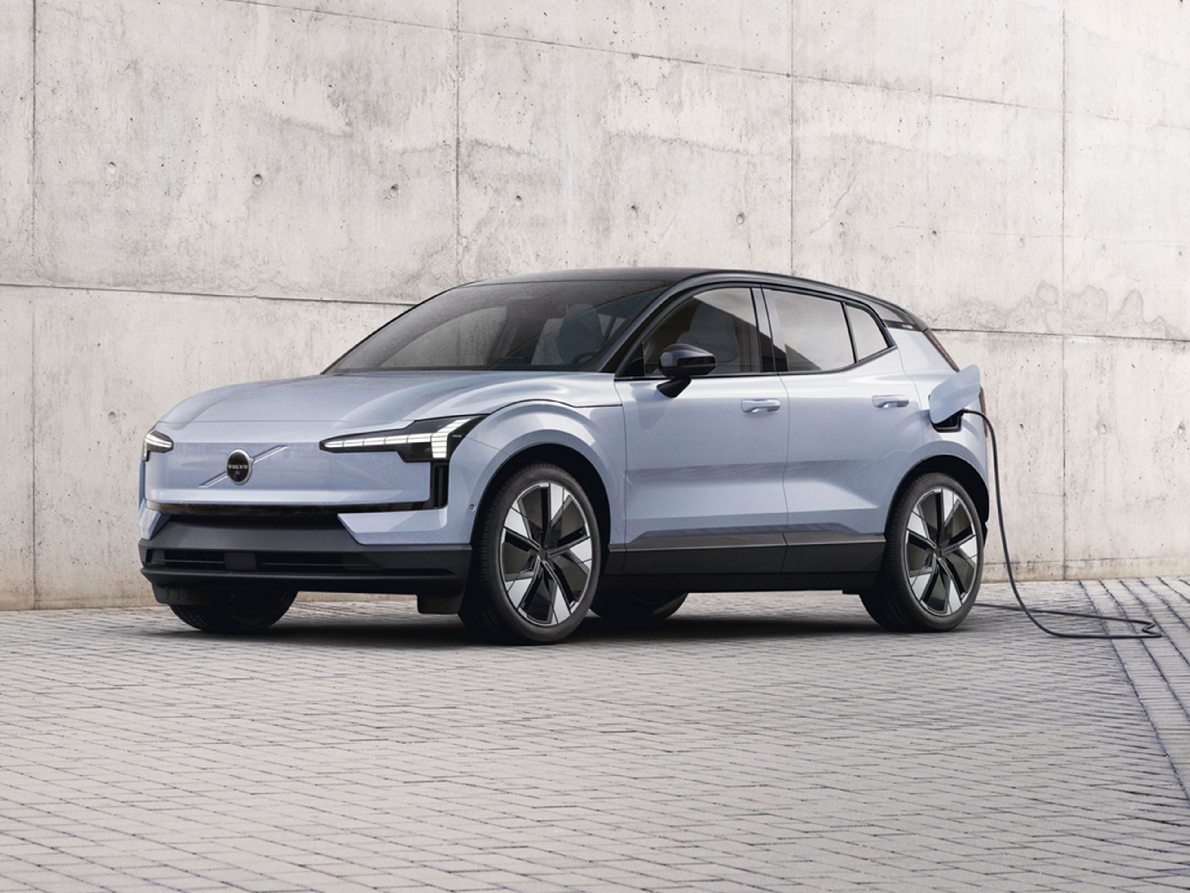 Lataukseen kytketty Cloud Blue värinen Volvo EX30 pysäköitynä vaalean betoniseinän eteen.