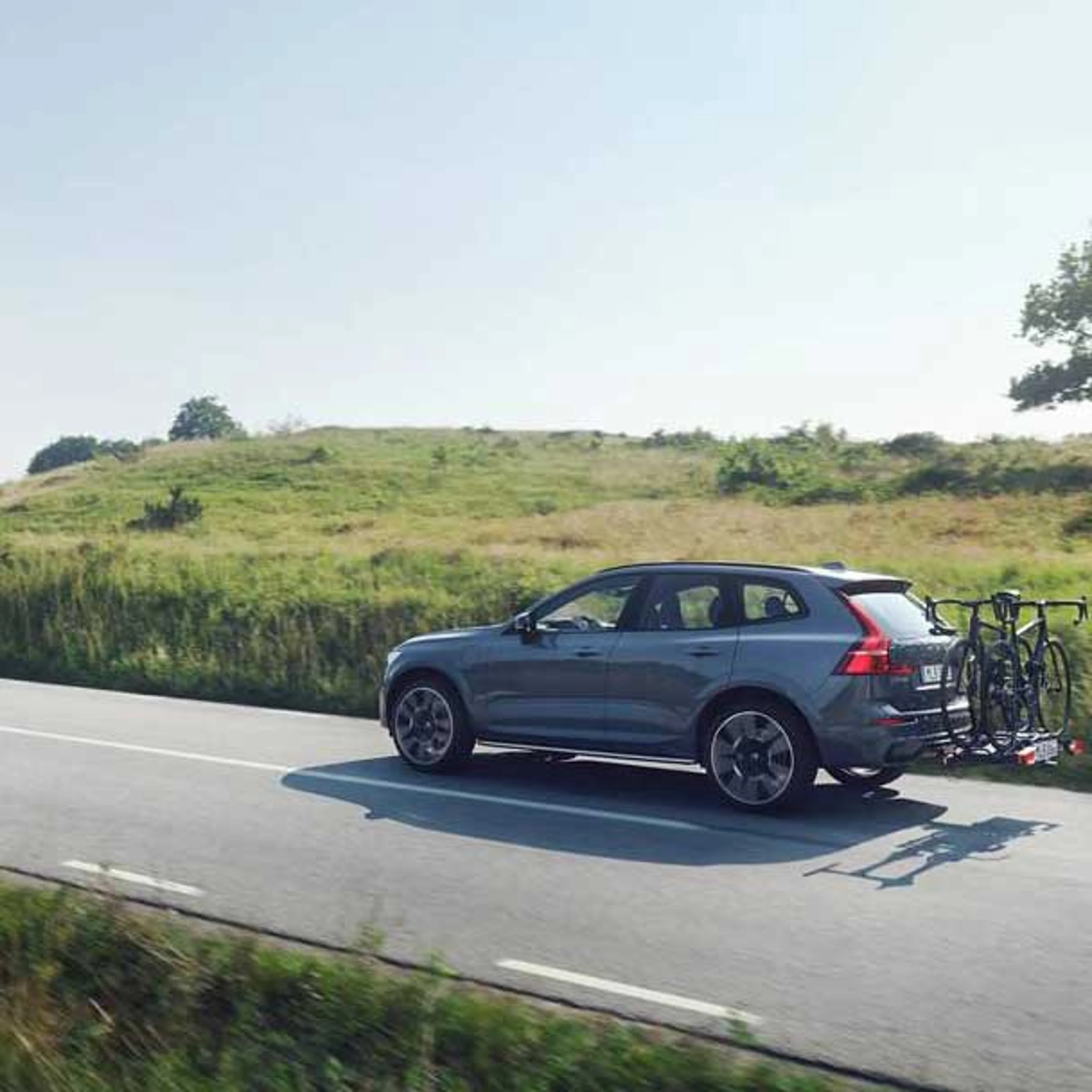 En grå Volvo XC60 SUV kører på en landevej med en cykelholder bagpå, der bærer to cykler. Scenen udspiller sig med grønne græsarealer og blå himmel i baggrunden.